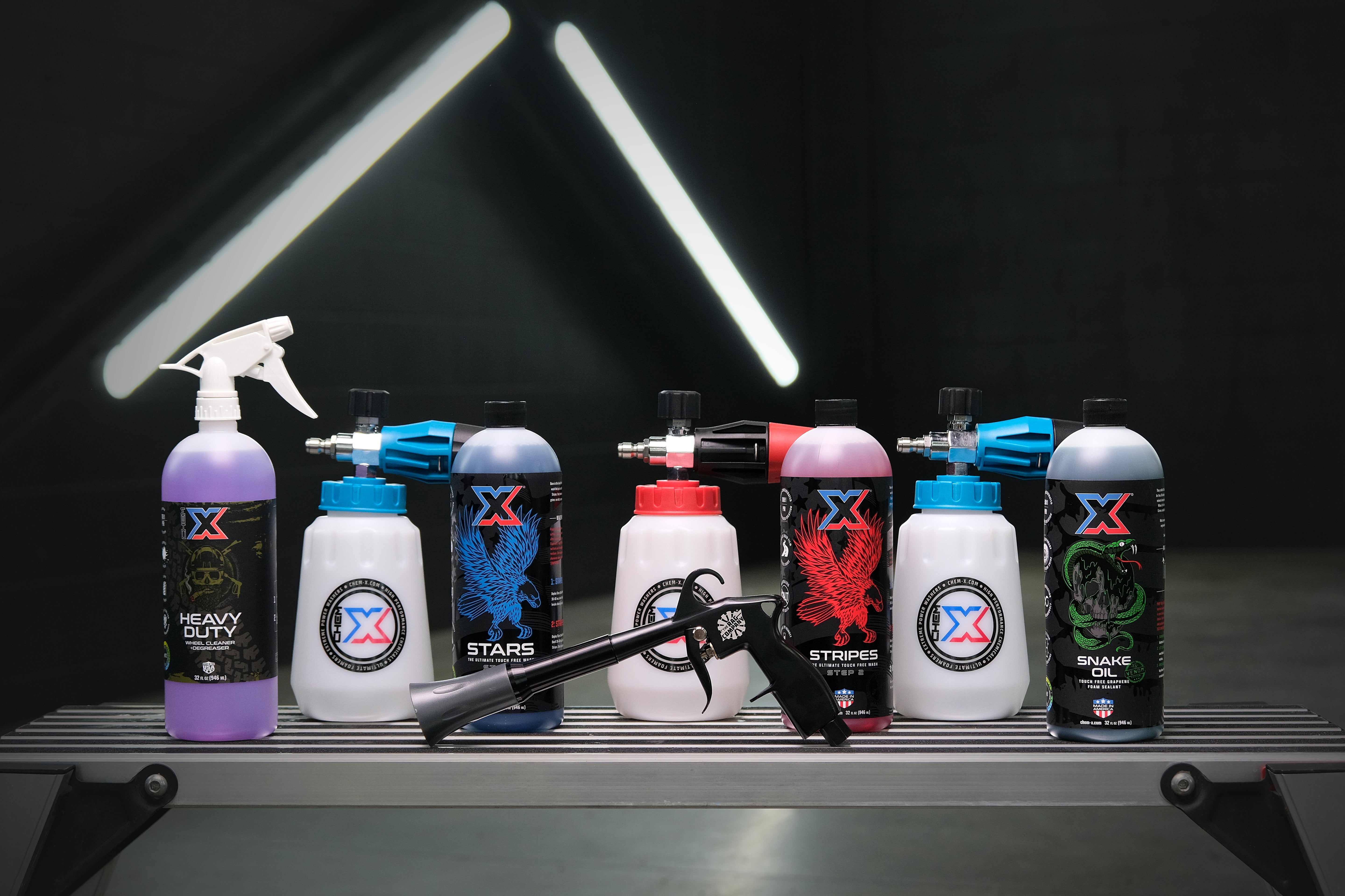 One 32 oz bottle of purple wheel cleaner and degreaser called 'Heavy Duty,' one 32 oz bottle of touch free soap called 'Stars,' one 32 oz bottle of touch free soap called 'Stripes,' and one 32 oz bottle of touch free graphene sealant called 'Snake Oil,' three foam cannons and an air blaster on a workbench in a wash bay setting with fluorescent lights.