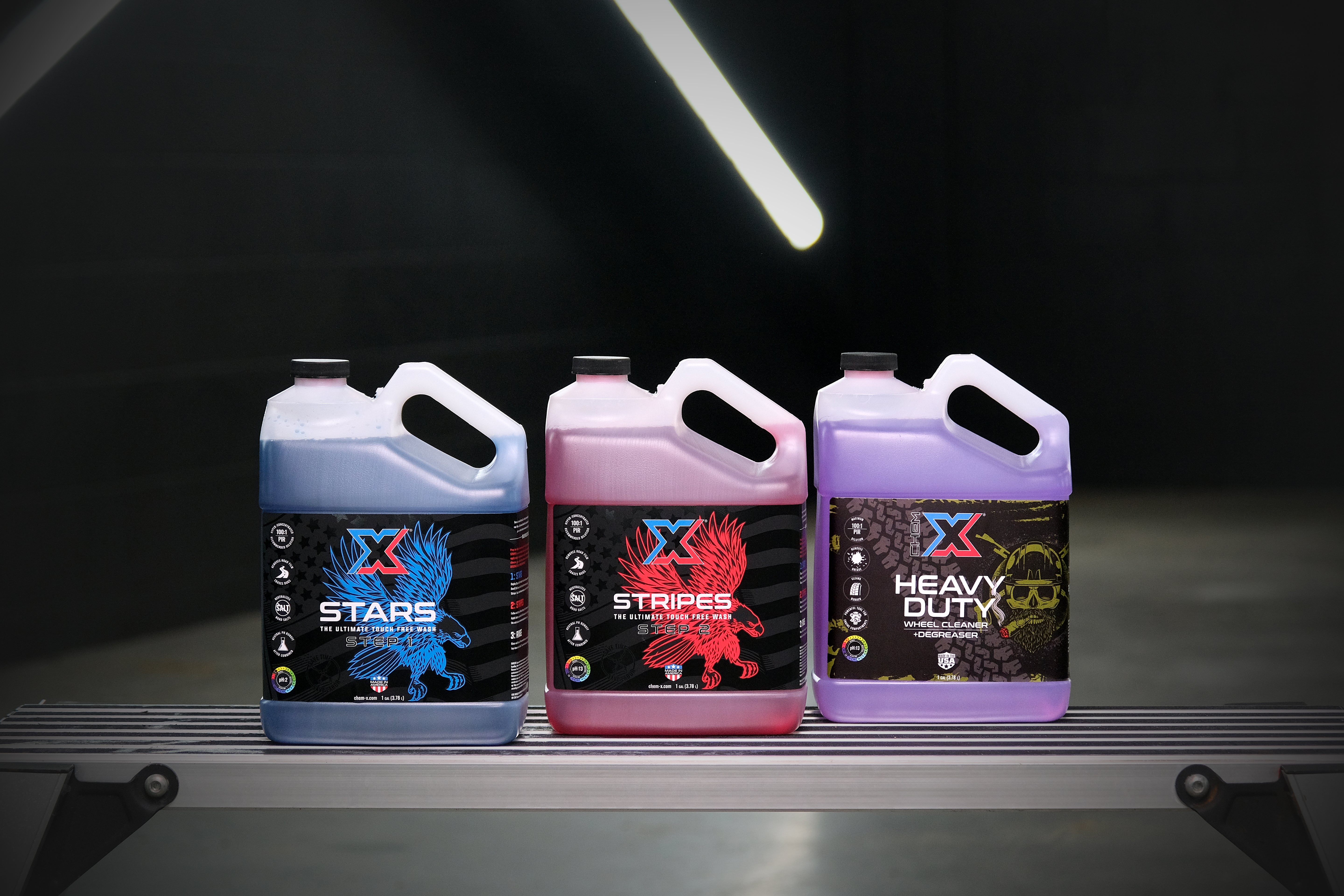 One gallon of blue touch free soap called 'Stars,' one gallon of red touch free soap called 'Stripes,' and one gallon of purple wheel cleaner and degreaser called 'Heavy Duty' on a dark background with fluorescent lights.