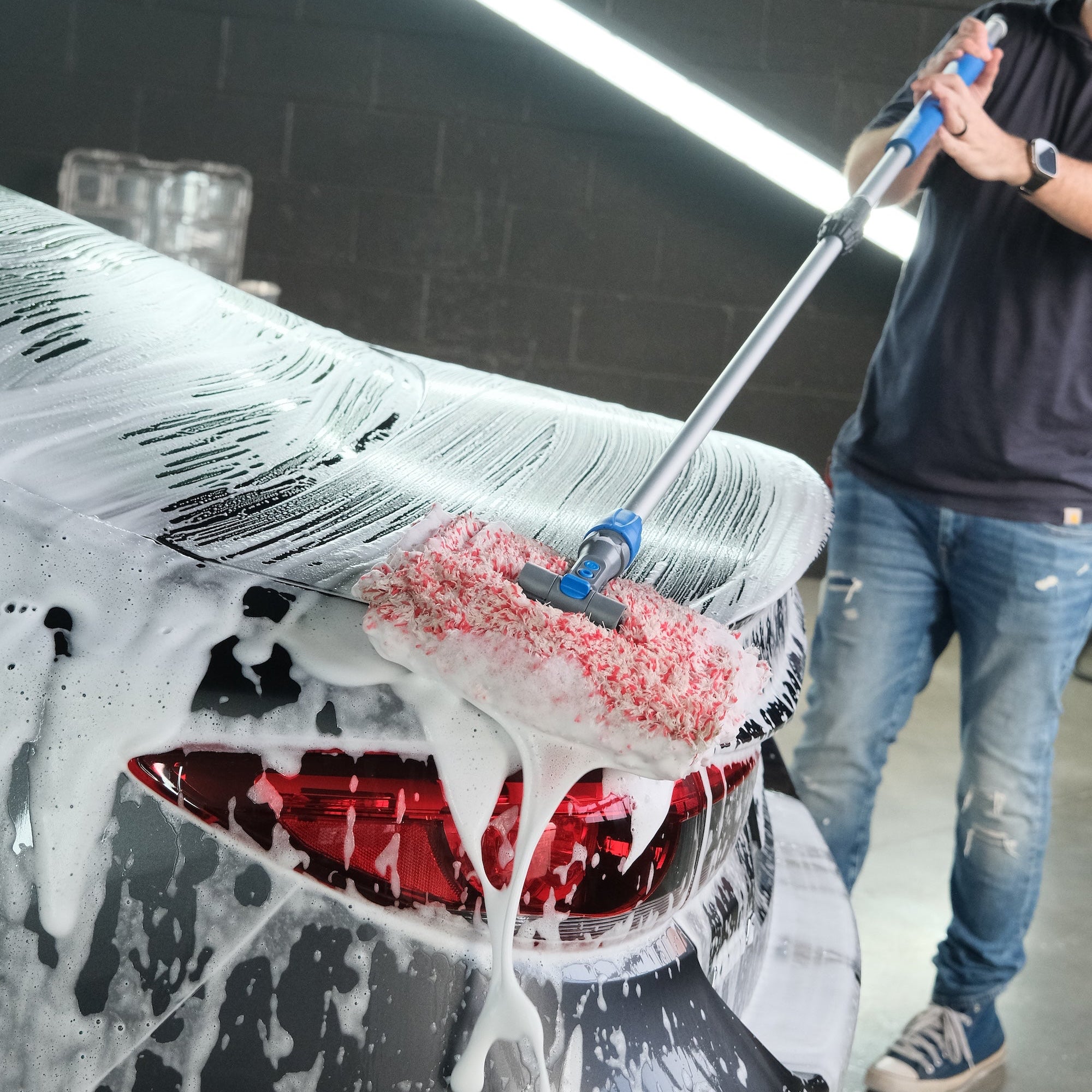 Person scrubbing a car down with foaming soap with a mitt on a stick in a wash bay setting with fluorescent lighting in the background.
