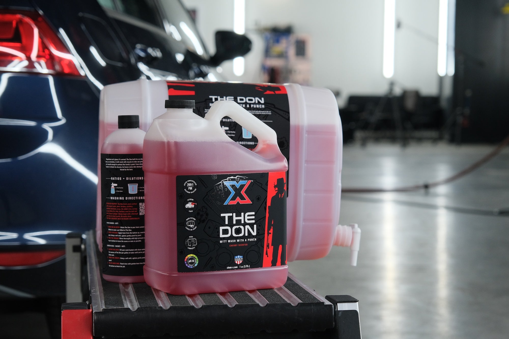 A one gallon bottle, 32 oz bottle, and 5 gallon pail of the same red vehicle mitt wash called 'The Don' lined up together on a workbench in front of a car in a wash bay setting.