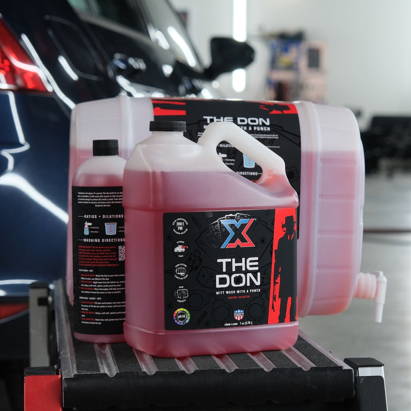 A one gallon bottle, 32 oz bottle, and 5 gallon pail of the same red vehicle mitt wash called 'The Don' lined up together on a workbench in front of a car in a wash bay setting.