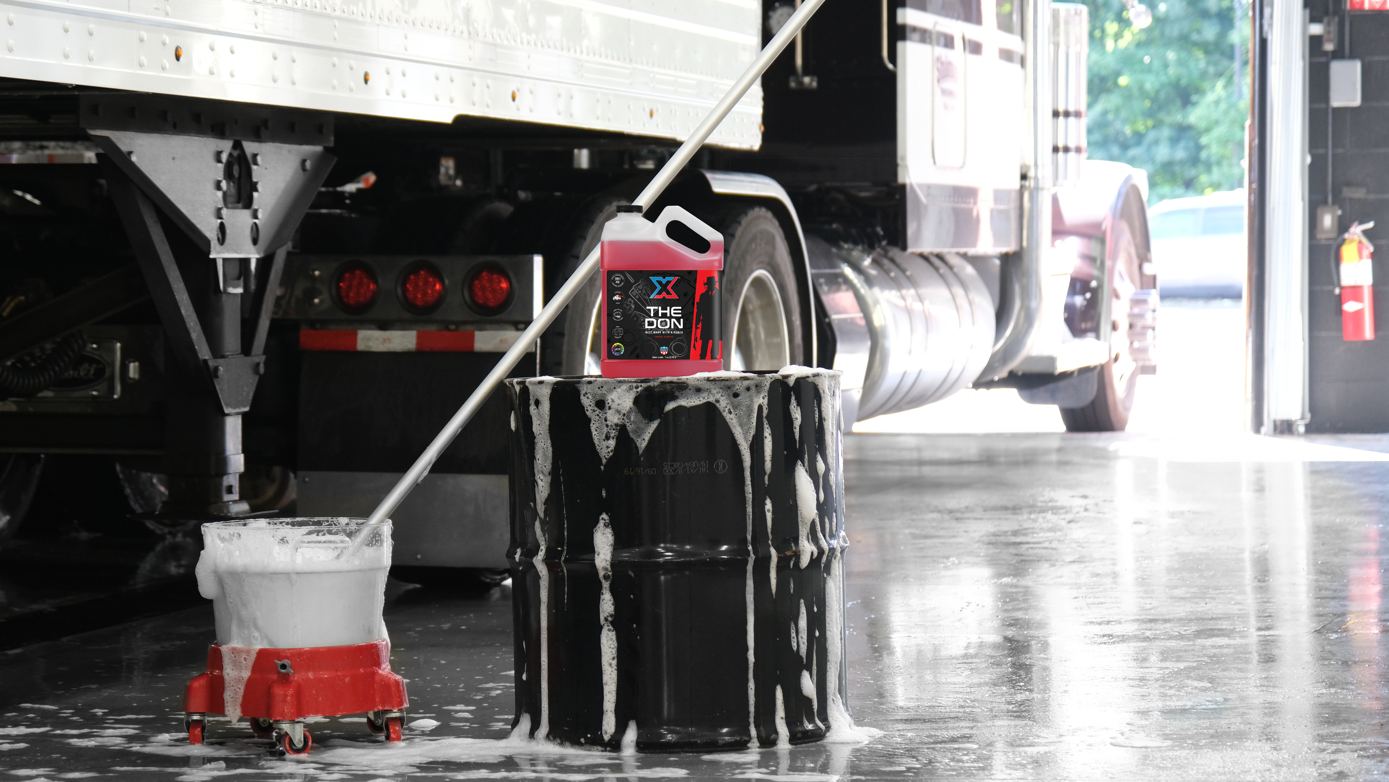 Black drum in front of a large truck with a gallon of red mitt wash called 'The Don' sitting on top of it and a mitt on a stick in a soapy bucket next to it in a wash bay setting.