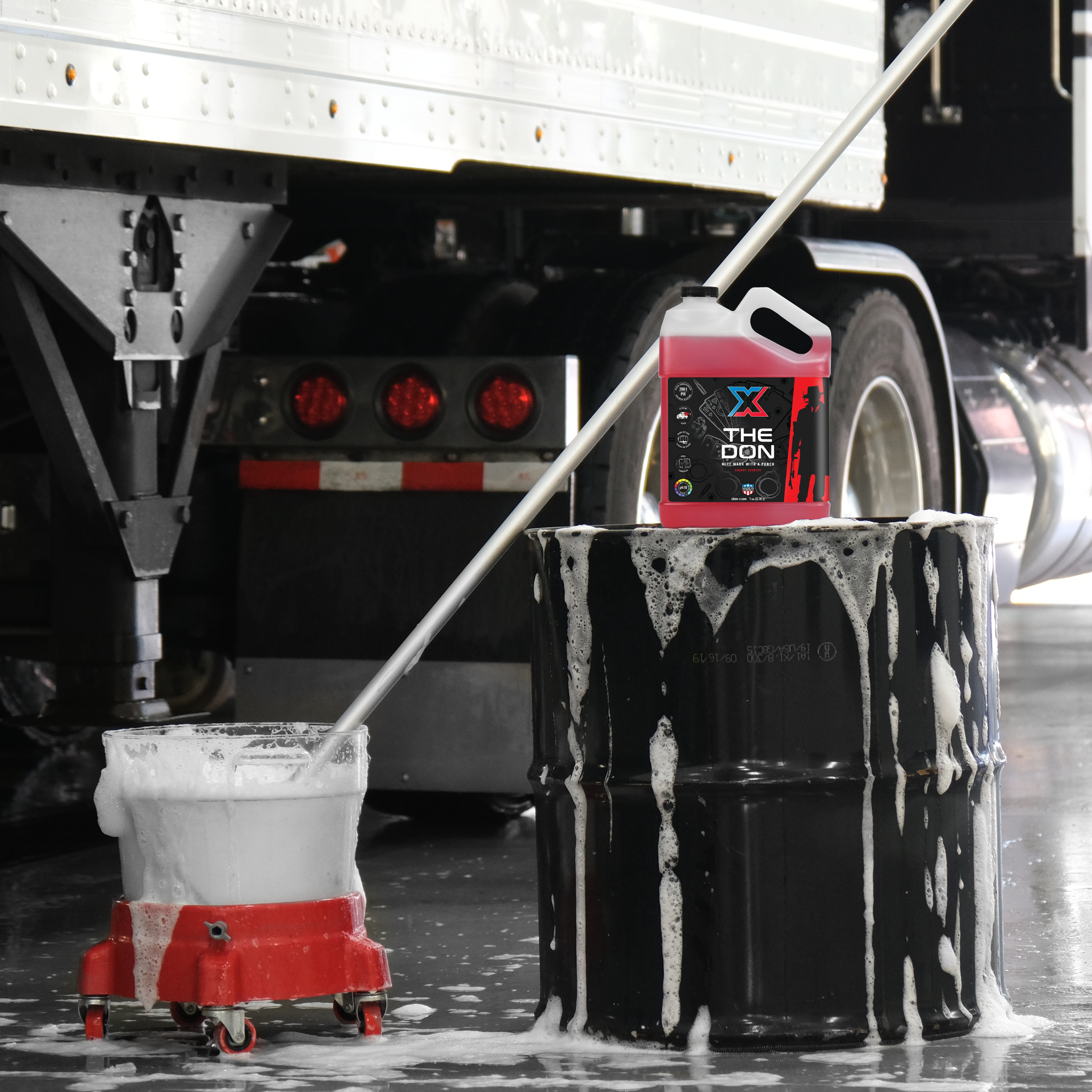 Black drum in front of a large truck with a gallon of red mitt wash called 'The Don' sitting on top of it and a mitt on a stick in a soapy bucket next to it in a wash bay setting.