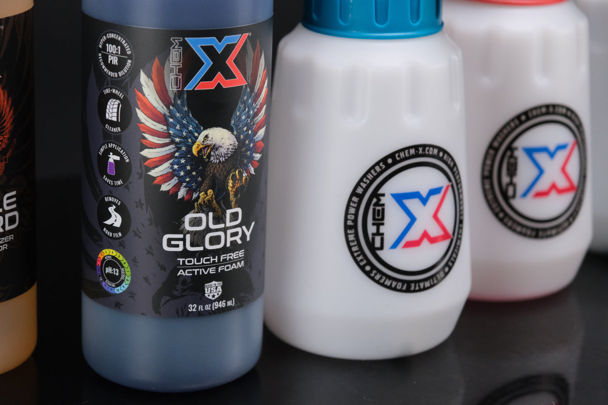 Bottle of black touch free one-step active foam vehicle wash with Chem-X branding on the label, next to two foam cannon bottles that are slightly out of focus with Chem-X branding on them, sitting on a black reflective surface.