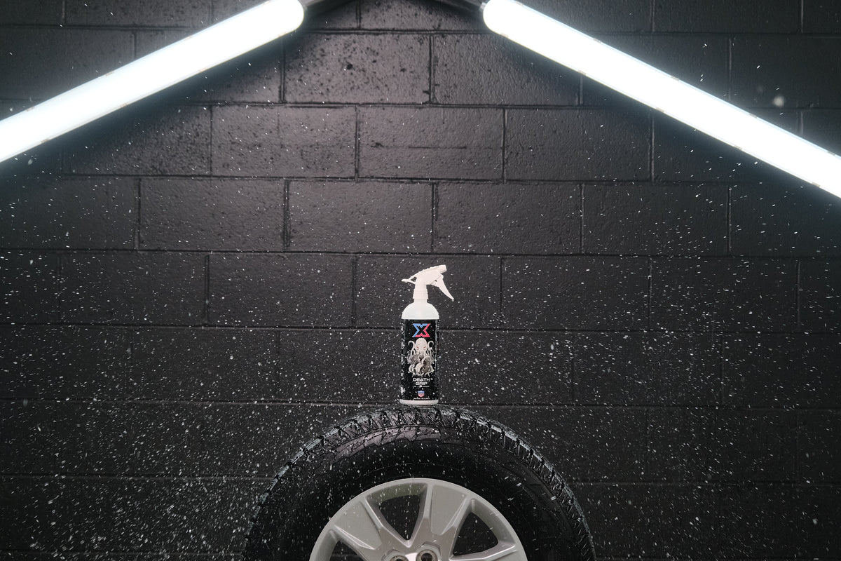 A bottle with a black label that has Chem-X branding on the label and a white trigger sprayer sitting on a tire with water droplets surrounding it in front of a black wall with two LED light strips.
