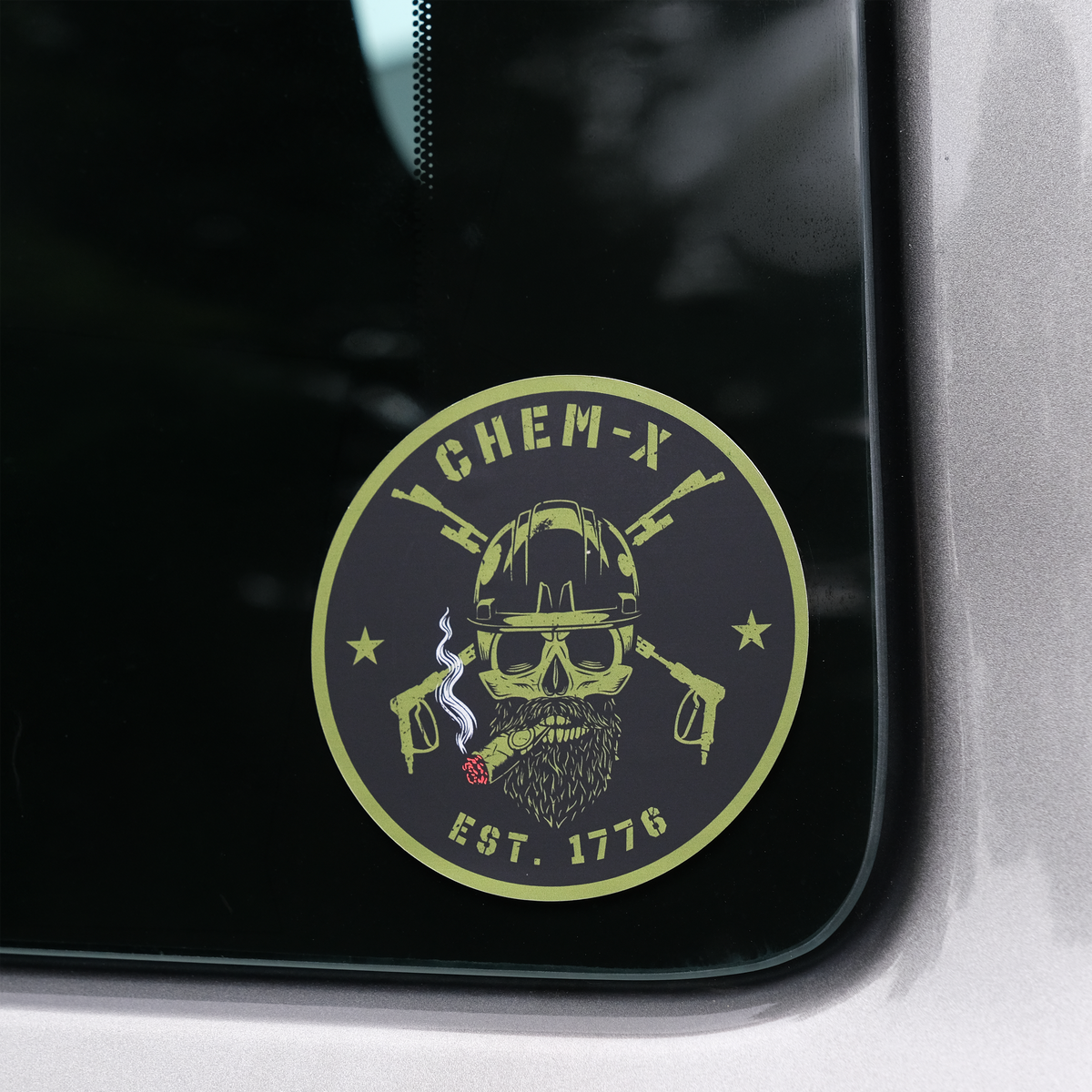 A circular black and green decal sticker featuring a skull with a hard hat, a beard, crossed lances, a burning cigar and the text 'CHEM-X EST. 1776', shown applied to a dark tinted truck window.