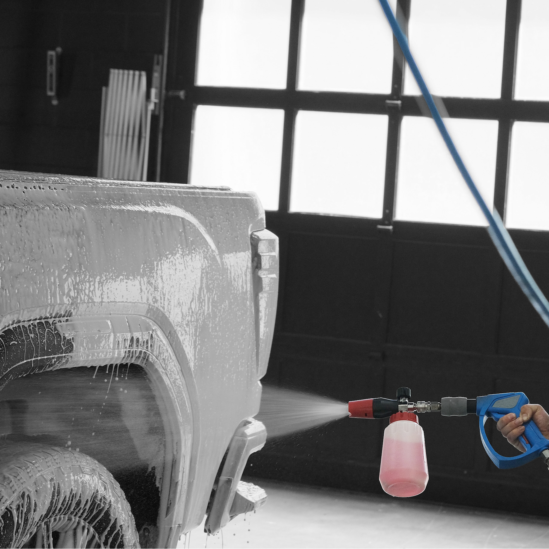 A close-up action shot of a person using a blue and gray MTM spray gun attached to a power washer to apply foam  onto the rear wheel area of a vehicle inside a garage.
