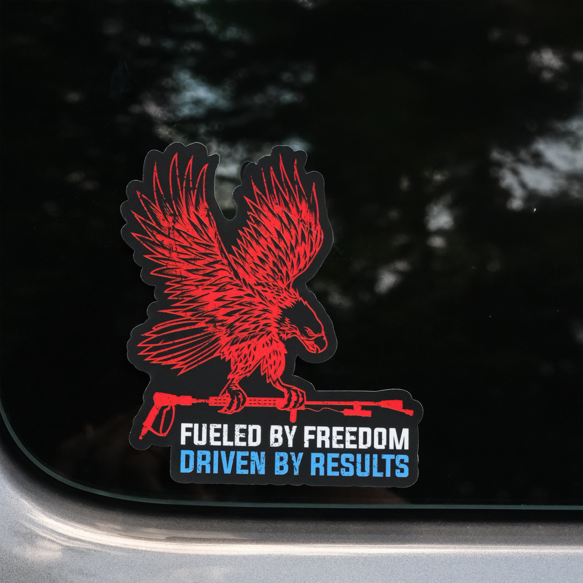 A die-cut vinyl sticker featuring a red eagle holding a rifle, with the text 'FUELED BY FREEDOM' in white and 'DRIVEN BY RESULTS' in blue, shown applied to a dark tinted car window.