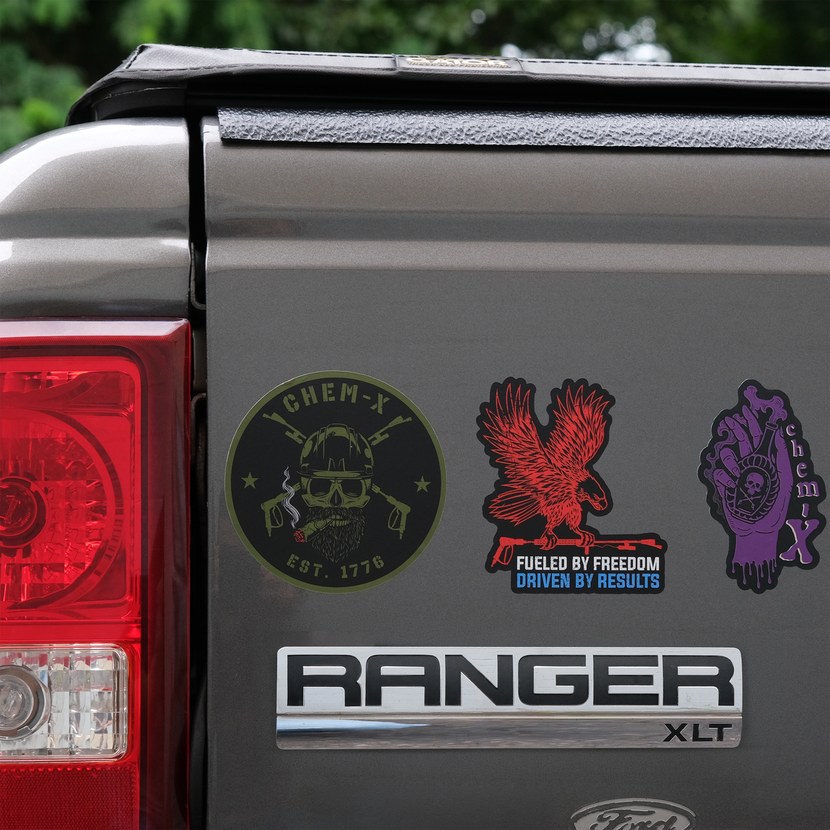 Three custom decals applied to the rear side of a gray Ford Ranger XLT truck bed. The decals include a green sticker with a skull smoking a cigar, a red sticker of an eagle carrying a lance with the words 'FUELED BY FREEDOM DRIVEN BY SUCCESS' underneath, and a purple sticker with a hand holding a potion with a skull on it on a red background.
