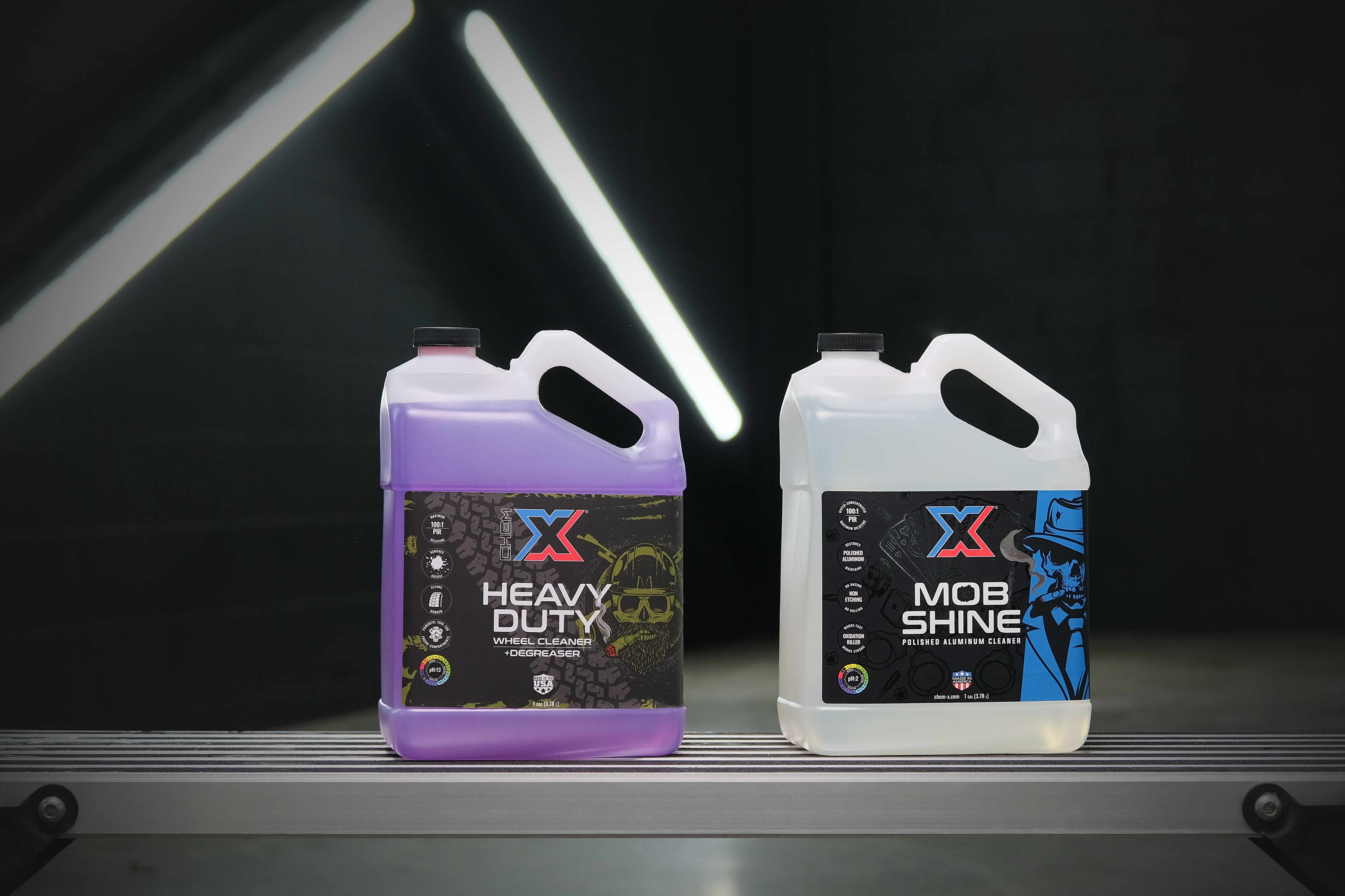 One gallon bottle of purple wheel cleaner and degreaser called 'Heavy Duty' sitting next to a one gallon bottle of clear polished aluminum cleaner called 'Mob Shine' on a workbench in a wash bay setting with fluorescent lighting in the background.