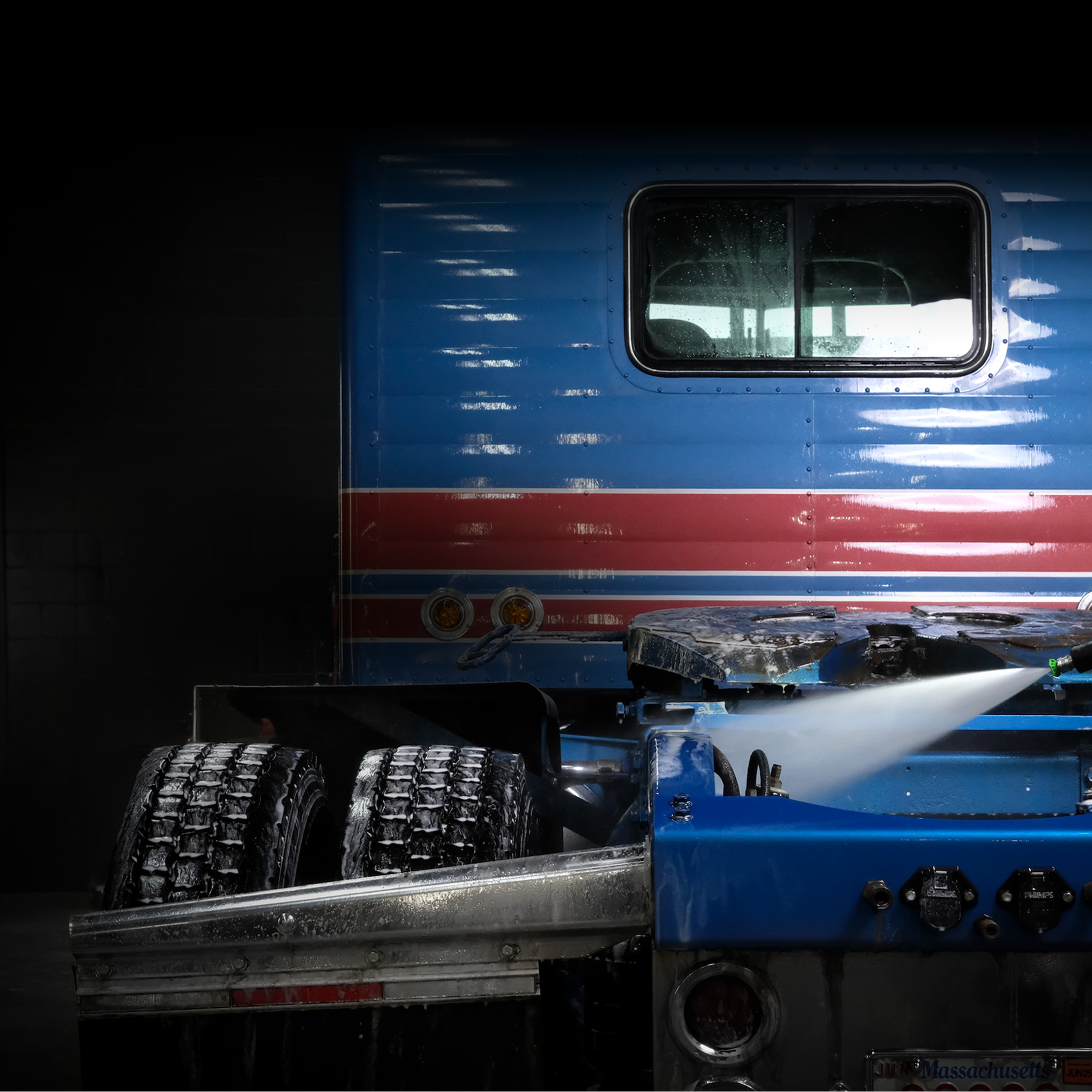 Close-up of a blue truck with a red stripe getting sprayed with power washer on dark background.