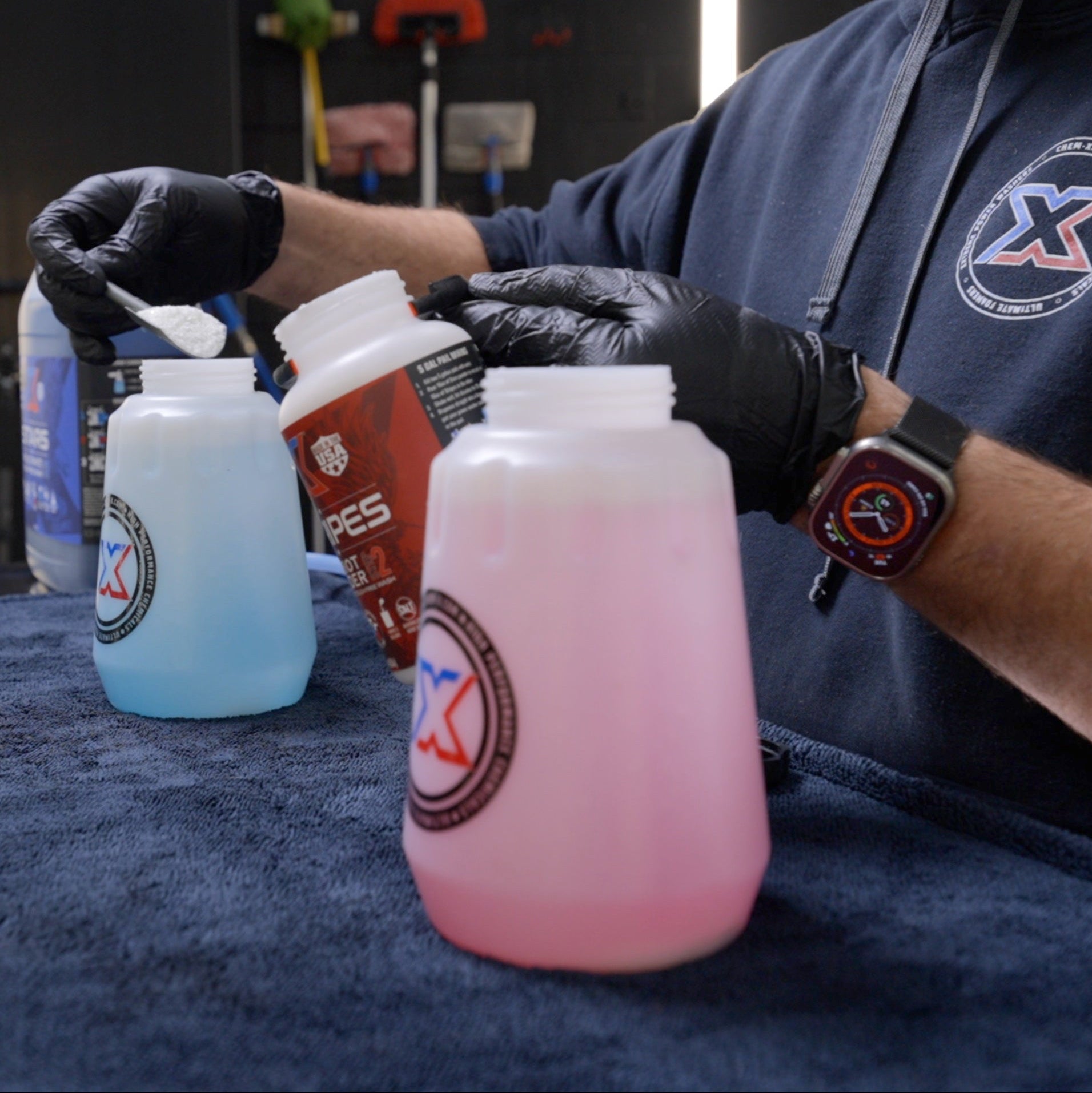 A person wearing black gloves and a black hoodie with a circular Chem-X logo scoops a white substance from a small bottle into a translucent plastic bottle filled with blue liquid. Another bottle with a red liquid sits in the foreground on a dark blue towel.