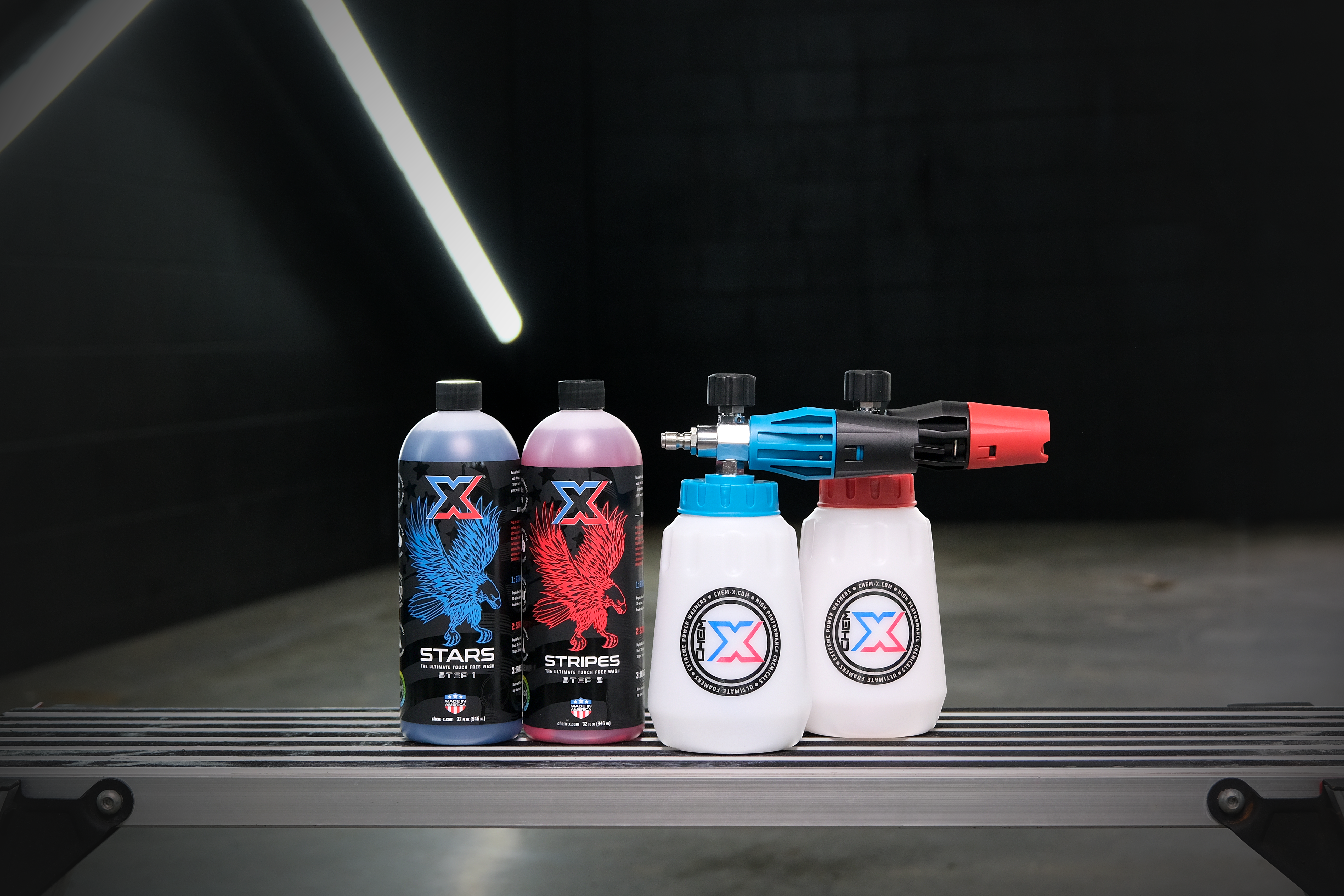One 32 oz bottle of blue liquid called 'Stars,' and one 32 oz bottle of red liquid called 'Stripes' sitting on a workbench next to a blue and a red foam cannon in a wash bay setting.