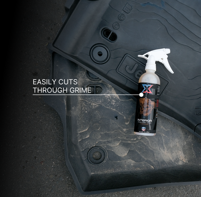 A bottle of orange leather-scented interior cleaner + detailer called Interior with a white tolco sprayer placed on a dirty black floor mat, with the text 'EASILY CUTS THROUGH GRIME' overlaid on the image.