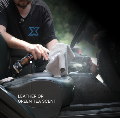 A person applies an orange leather-scented interior cleaner + detailer called Interior from a spray bottle onto a gray cloth with text overlay reading 'LEATHER OR GREEN TEA SCENT.'
