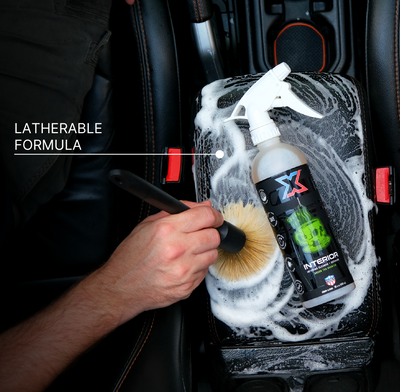 A hand uses a boar's hair brush to clean a car center console foam-covered surface with a bottle with orange green tea-scented interior cleaner + detailer called Interior laying across the center console. Text on the left reads 'LATHERABLE FORMULA'.