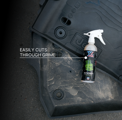 A bottle of orange green tea-scented interior cleaner + detailer called Interior with a white tolco sprayer placed on a dirty black floor mat, with the text 'EASILY CUTS THROUGH GRIME' overlaid on the image.