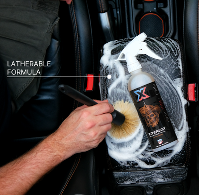 A hand uses a boar's hair brush to clean a car center console foam-covered surface with a bottle with orange leather-scented interior cleaner + detailer called Interior laying across the center console. Text on the left reads 'LATHERABLE FORMULA'.
