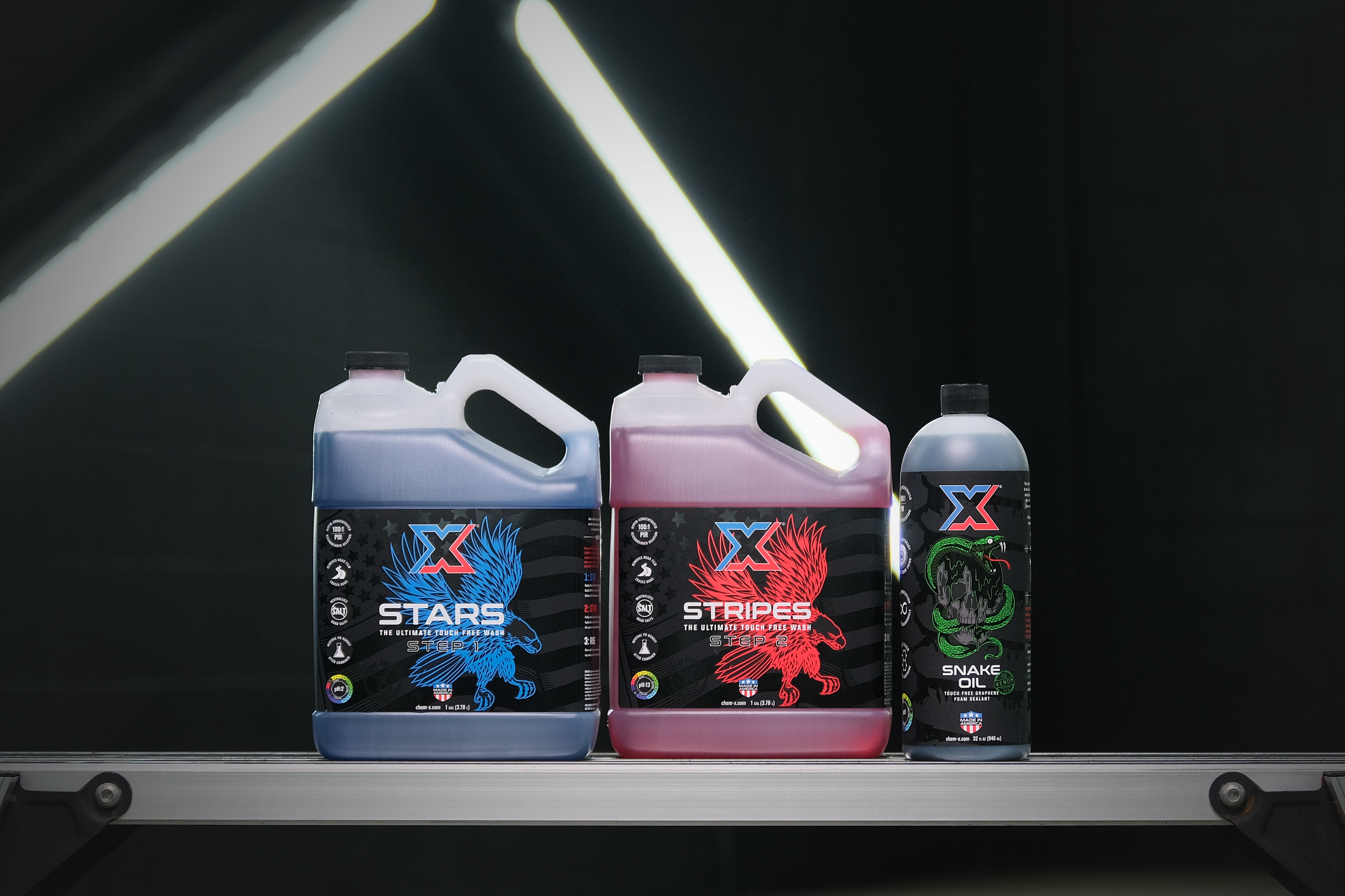 Three bottles of vehicle cleaning products with different labels and different colored liquids sitting on a workbench in front of a black wall with fluorescent lights.