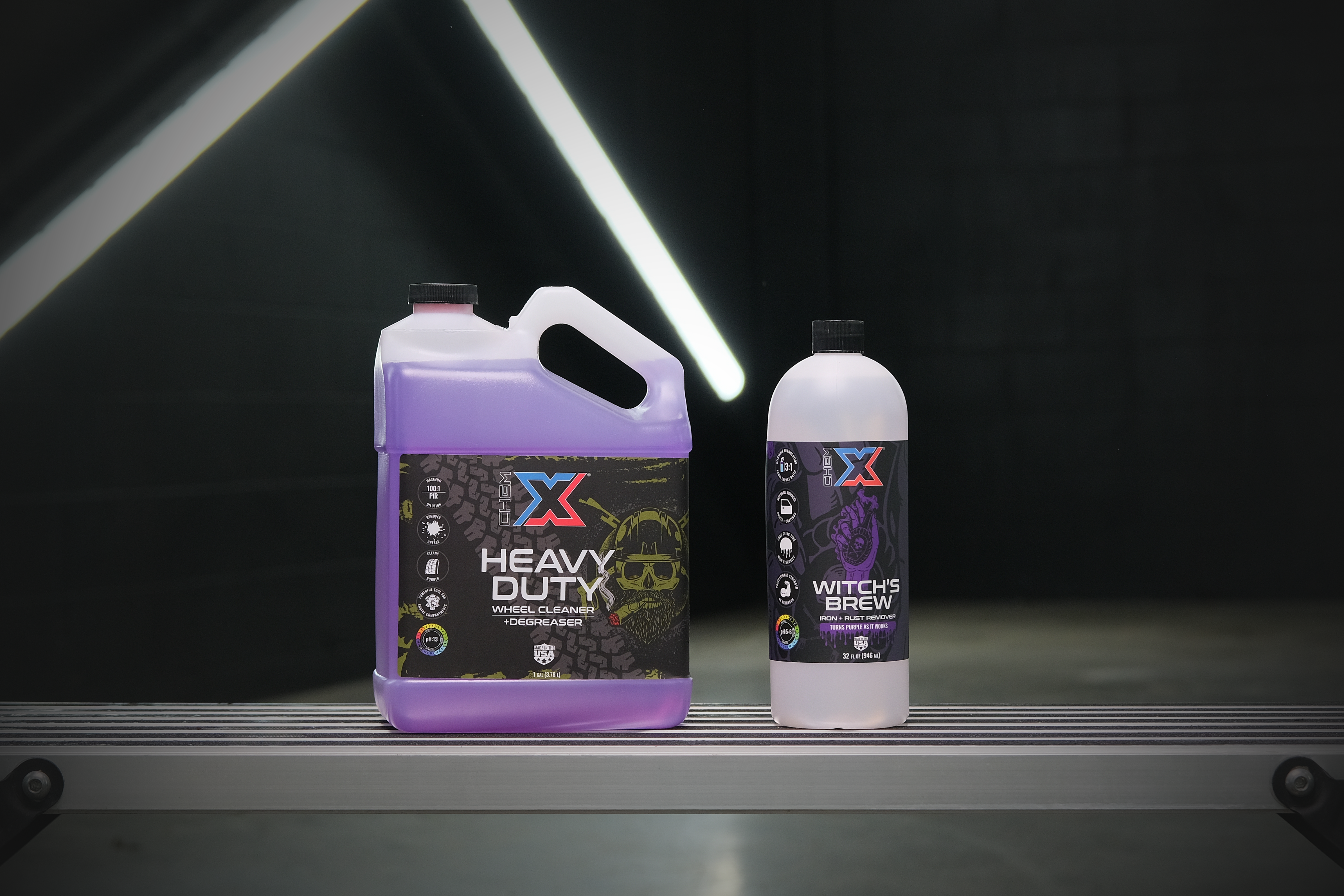 One gallon bottle of purple wheel cleaner and degreaser called 'Heavy Duty' next to a 32 oz bottle of clear iron and rust remover called 'Witch's Brew' on a workbench in a wash bay setting with fluorescent lighting in the background.