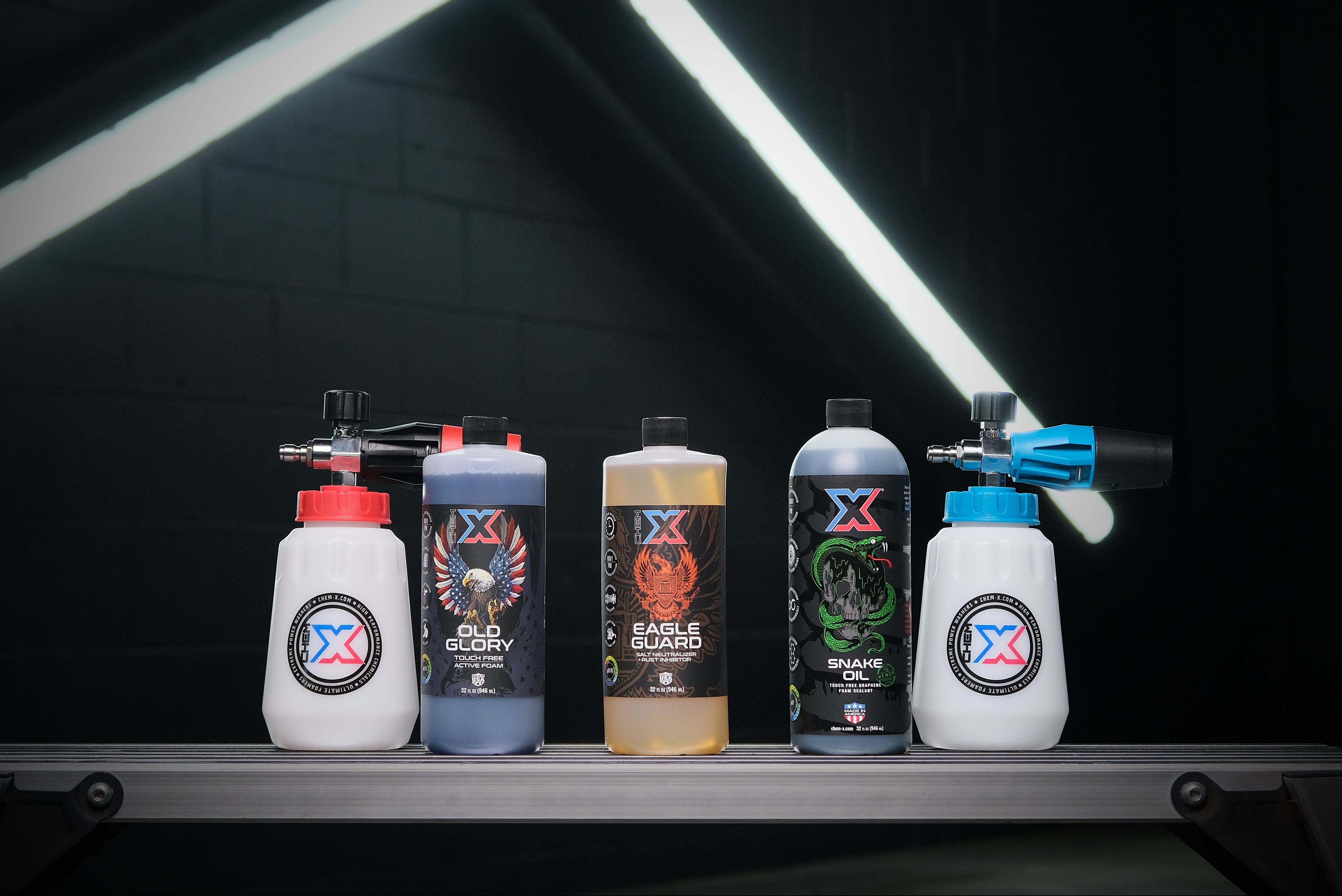 One foam cannon with a red cap lined up next to one 32 oz bottle of black touch free active foam vehicle wash called 'Old Glory,' one 32 oz bottle of orange salt neutralizer and rust inhibitor called 'Eagle Guard,' one 32 oz bottle of green touch free graphene foam sealant called 'Snake Oil,' and one foam cannon with a blue cap on a workbench in a dark wash bay setting with fluorescent lighting in the background.