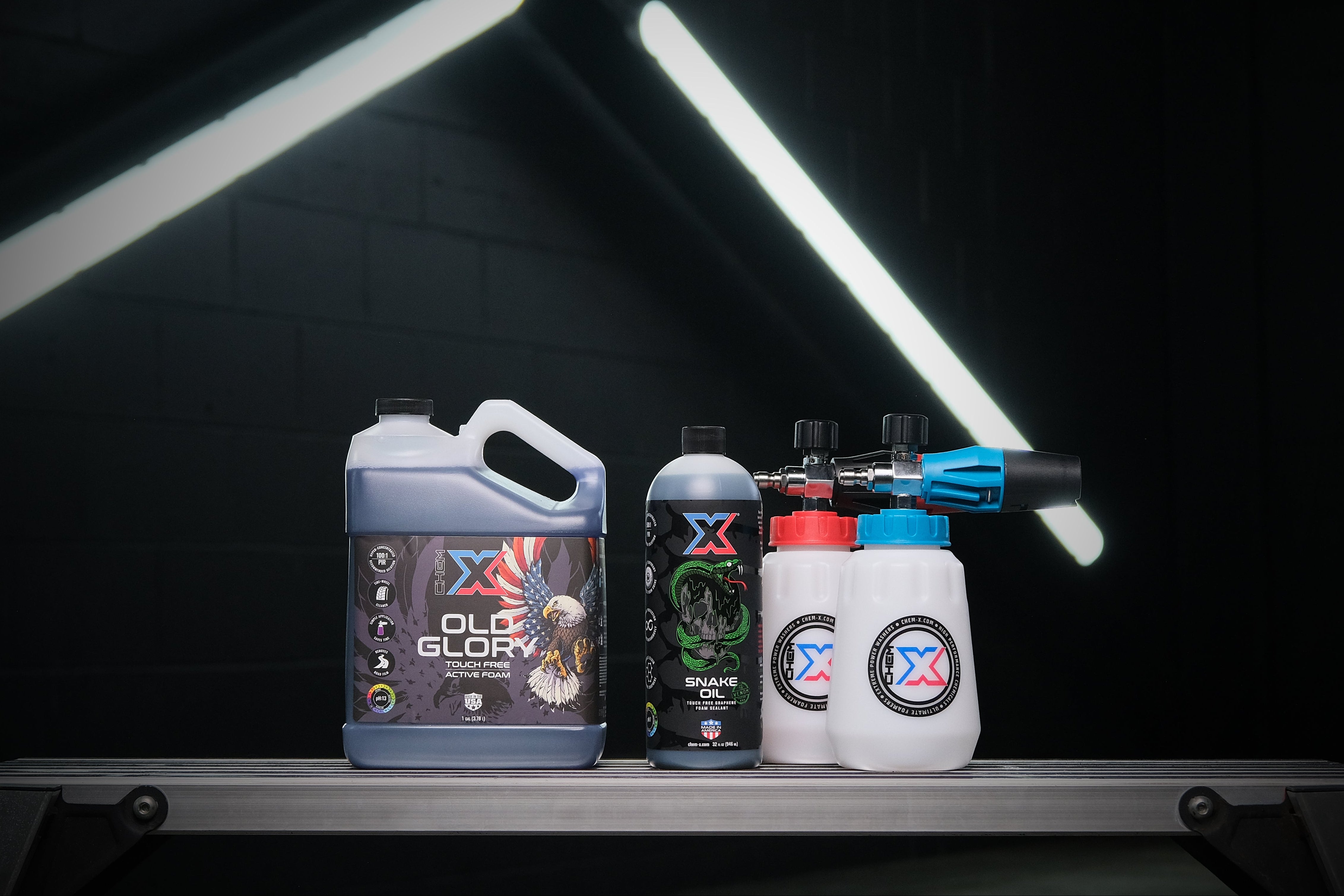 One gallon of black touch free active foam vehicle wash called 'Old Glory' and a 32 oz green touch free graphene foam sealant called 'Snake Oil' sitting on a workbench in a wash bay setting next to a foam cannon with a blue top and a foam cannon with a red top with fluorescent lights in the background.