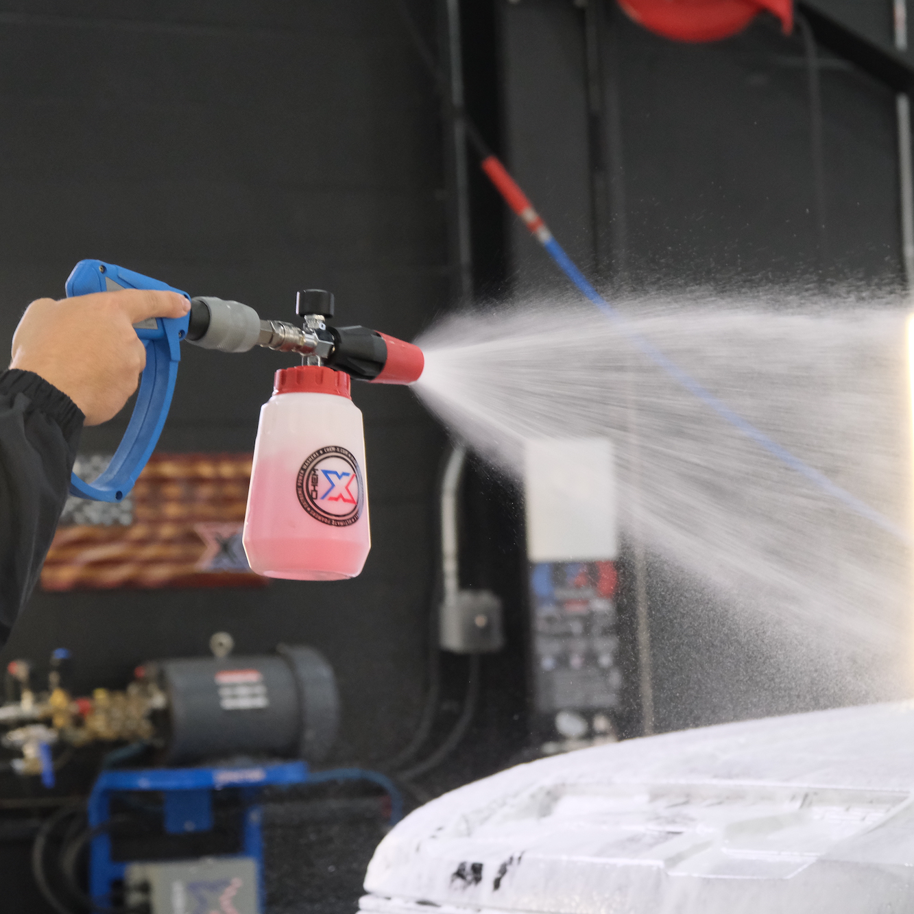 A person holding a blue and gray MTM spray gun attached to a power washer spraying a red touch-free vehicle wash called Stripes onto a vehicle in a workshop setting with a dark background.