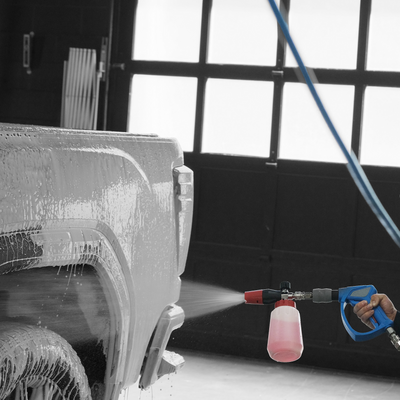 A close-up action shot of a person using a blue and gray MTM spray gun attached to a power washer to apply foam  onto the rear wheel area of a vehicle inside a garage.