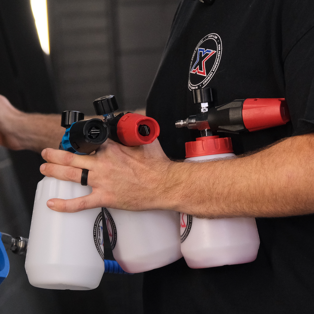 A person wearing a black Chem-X branded shirt holds three white foam cannon bottles with red and blue spray nozzles attached. The person's hands, forearms and torso are visible against a dark background.