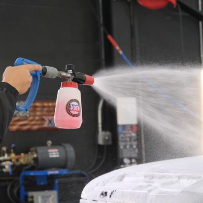 A person holding a blue and gray MTM spray gun attached to a power washer spraying a red touch-free vehicle wash called Stripes onto a vehicle in a workshop setting with a dark background.