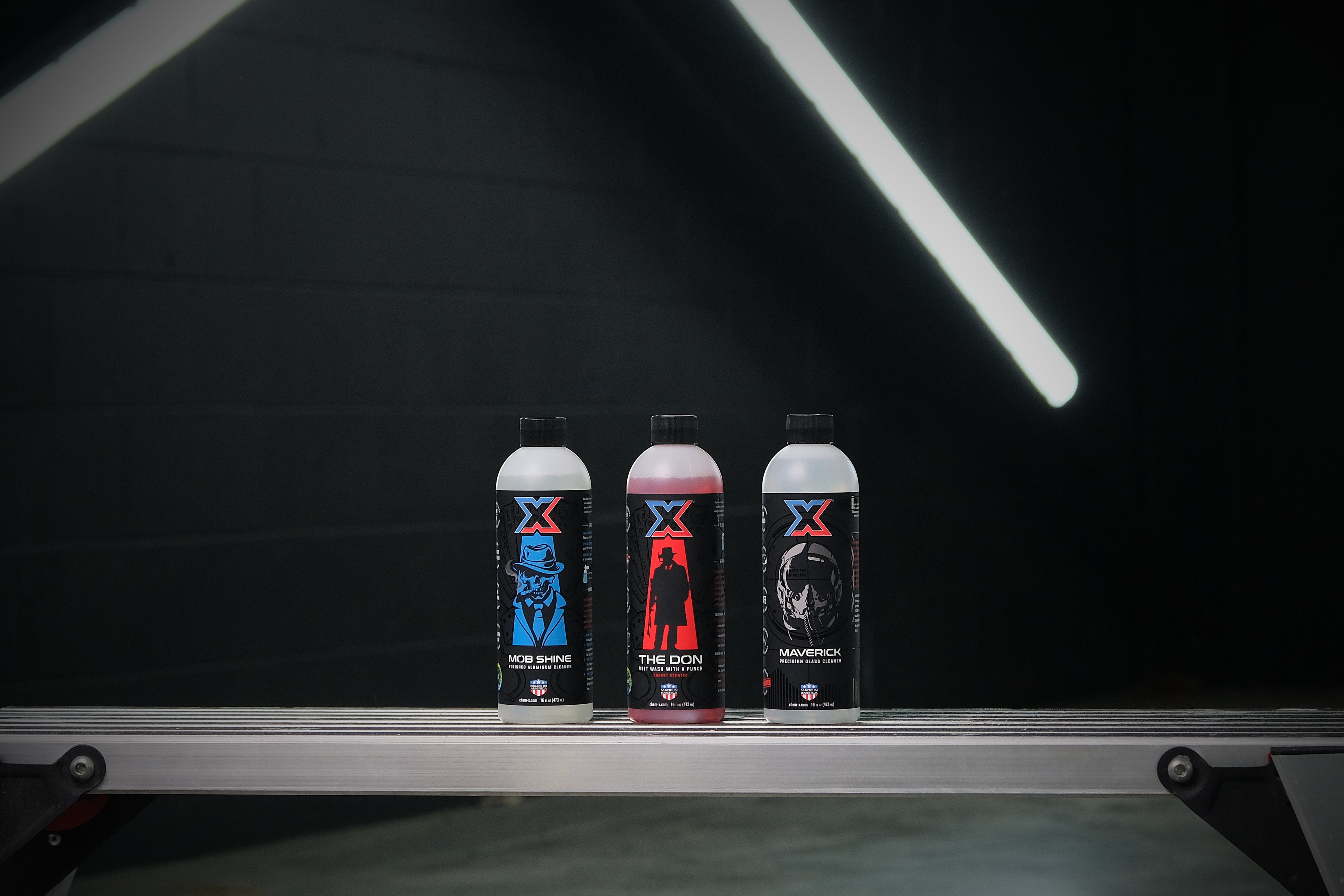 One 16 oz bottle of clear polished aluminum cleaner called 'Mob Shine,' one 16 oz bottle of red mitt wash called 'The Don,' and one 16 oz bottle of clear precision glass cleaner called 'Maverick' lined up on a workbench in a dark wash bay setting with fluorescent lighting behind.