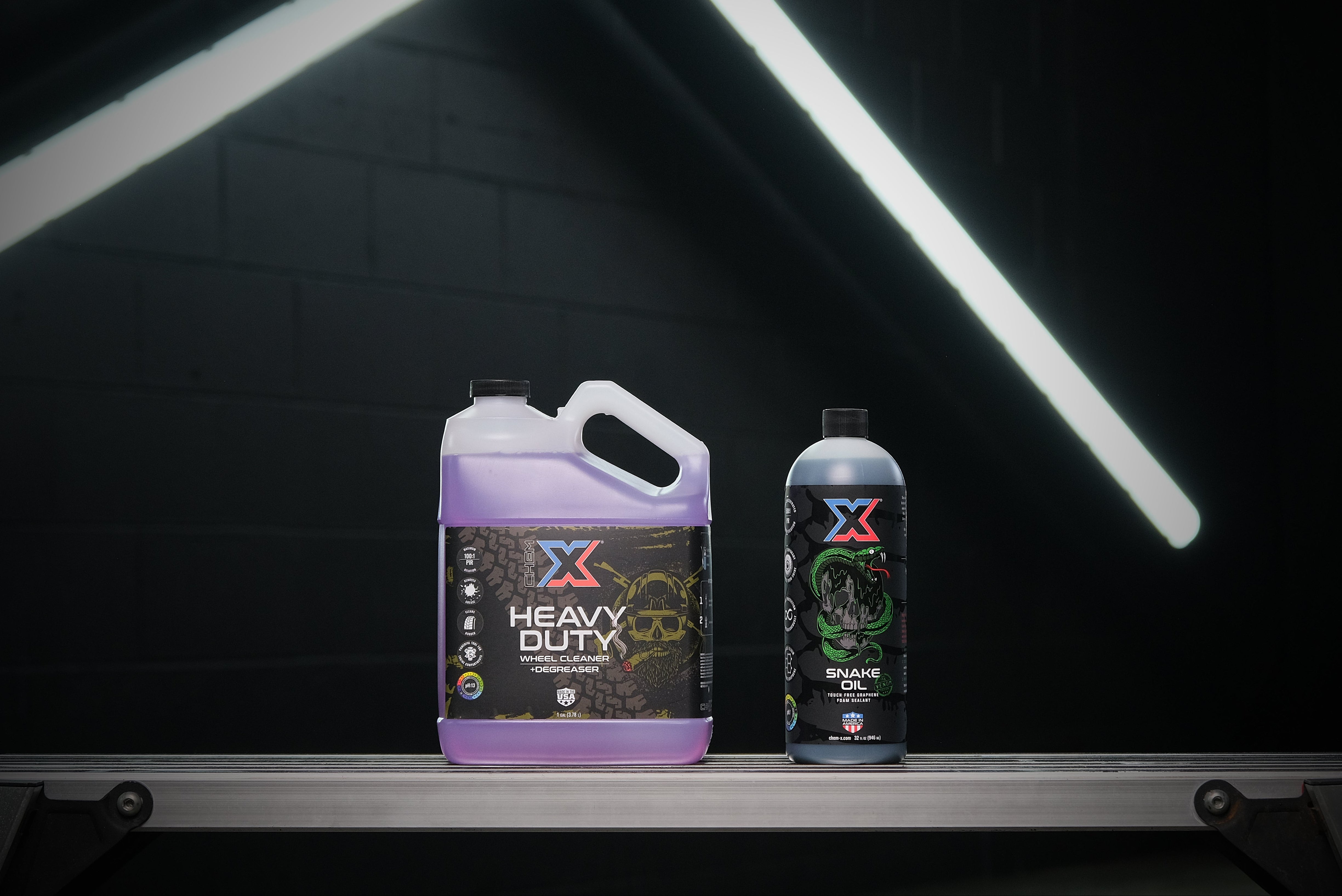 One gallon bottle of purple wheel cleaner and degreaser called 'Heavy Duty' sitting next to a 32 oz bottle of green touch free graphene foam sealant called 'Snake Oil' on a workbench in a dark wash bay setting with fluorescent lighting in the background.