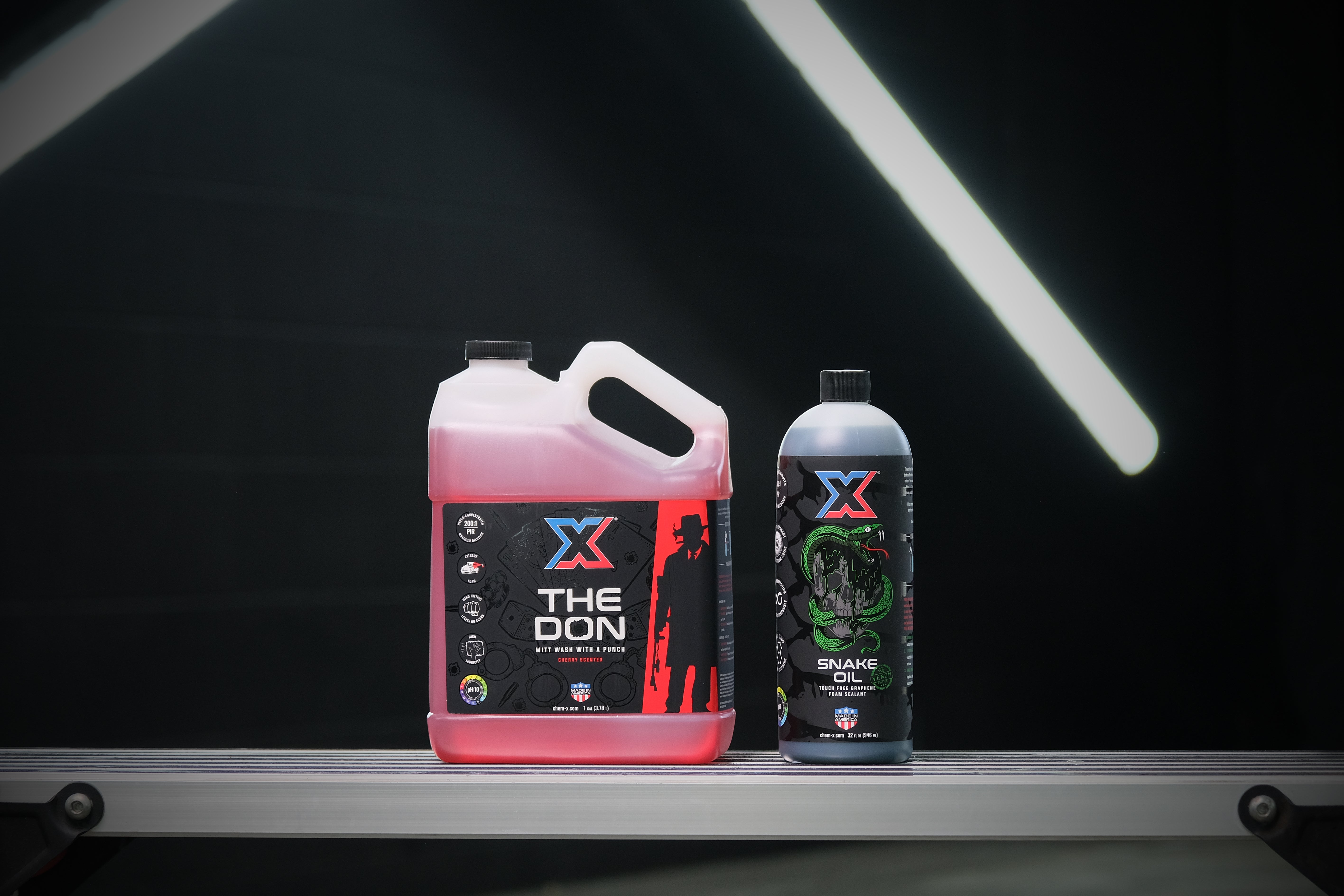 One gallon bottle of red mitt wash called 'The Don' and one 32 oz bottle of green touch free graphene foam sealant called 'Snake Oil' sitting on a work bench in a dark wash bay setting with fluorescent lighting in the background.