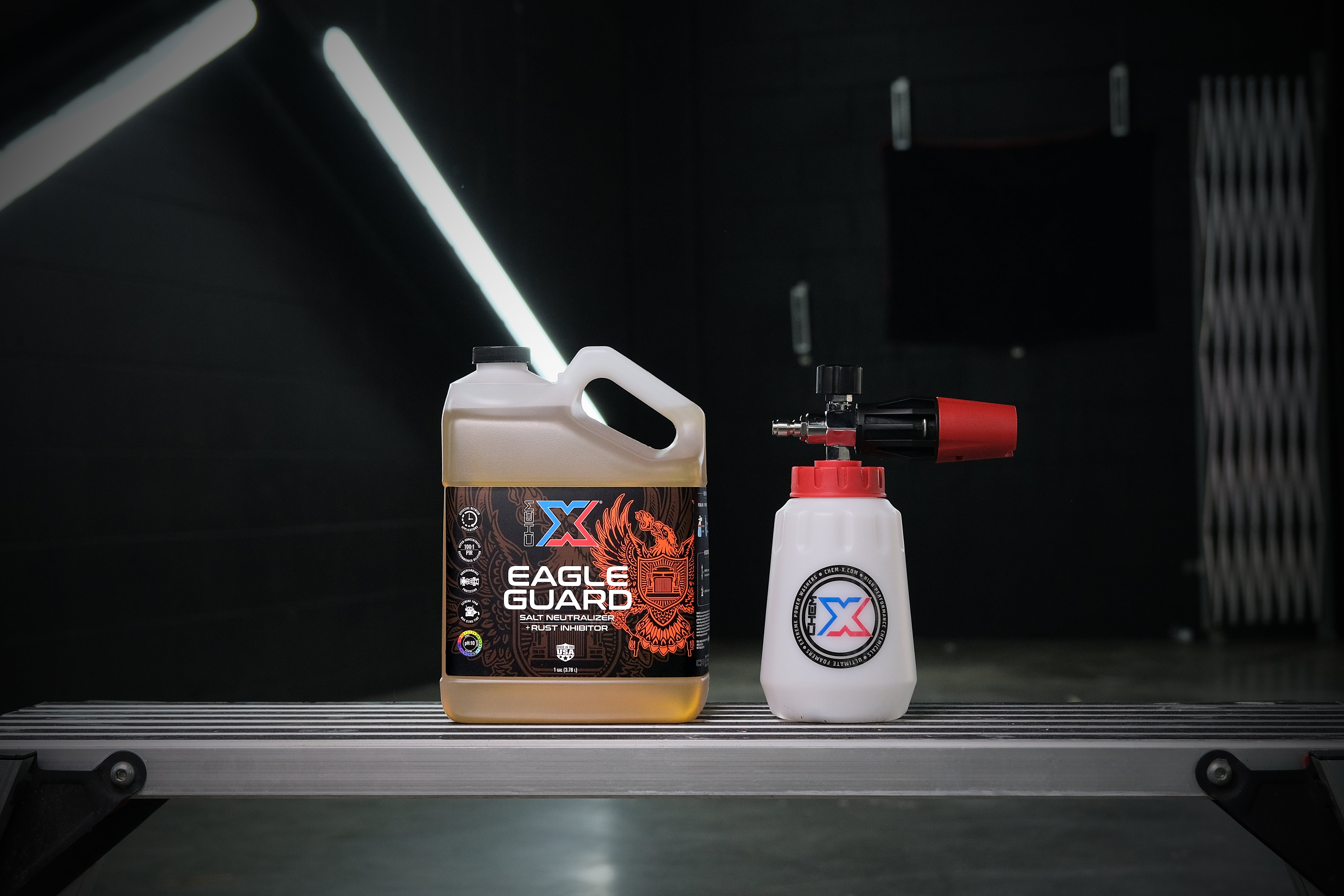 One gallon bottle of orange salt neutralizer and rust inhibitor called 'Eagle Guard' sitting next to a foam cannon with a red cap on a workbench in a dark wash bay setting with fluorescent lights in the background.