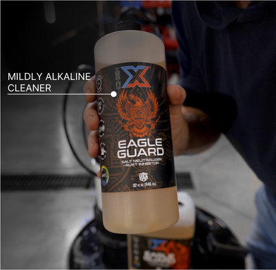 A hand holds a bottle of Chem-X Eagle Guard salt neutralizer and rust inhibitor with the text saying 'MILDLY ALKALINE CLEANER' overlaid across the top left of the image. The bottle has a black label with an orange eagle graphic and white text, and the liquid inside is a pale orange color.