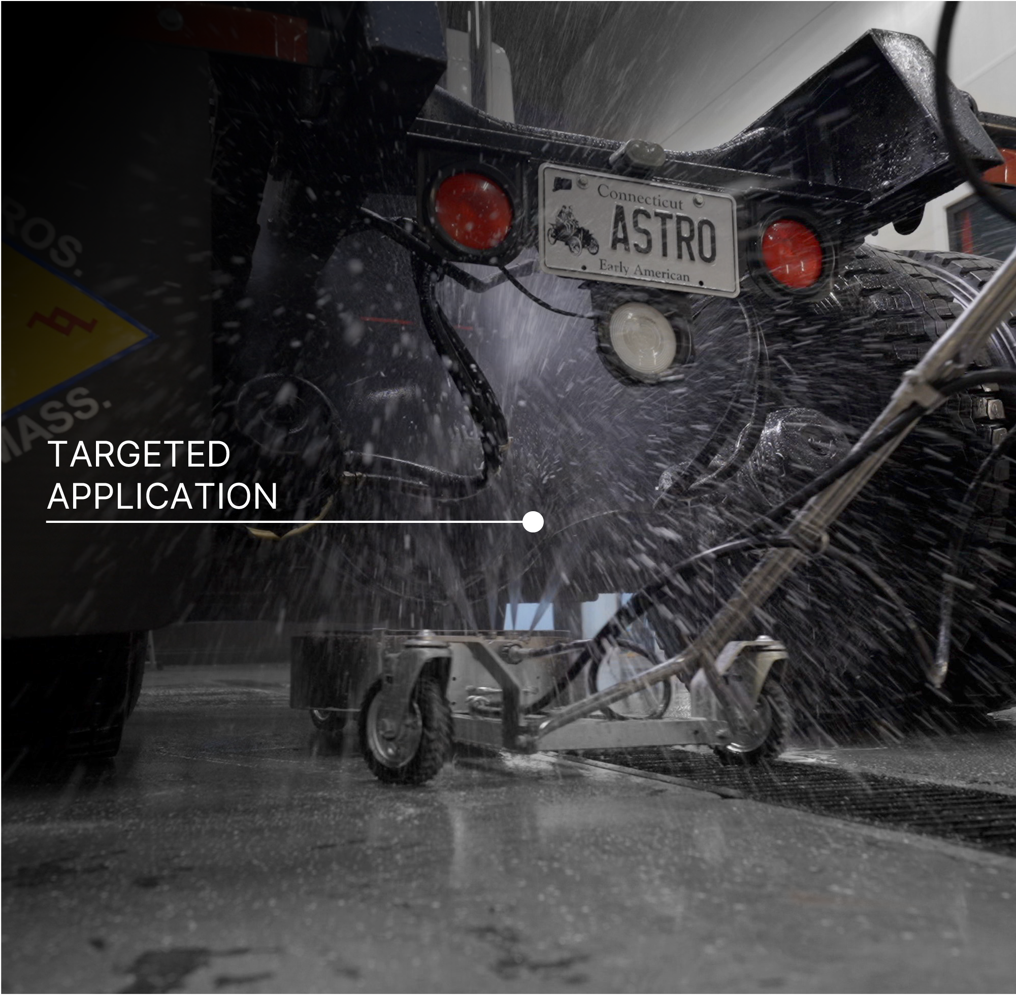 A close-up action shot of a high-pressure undercarriage cleaner being used to wash a black 18-wheeler, with water spraying forcefully. There is text in the middle left of the image that reads 'TARGETED APPLICATION.'