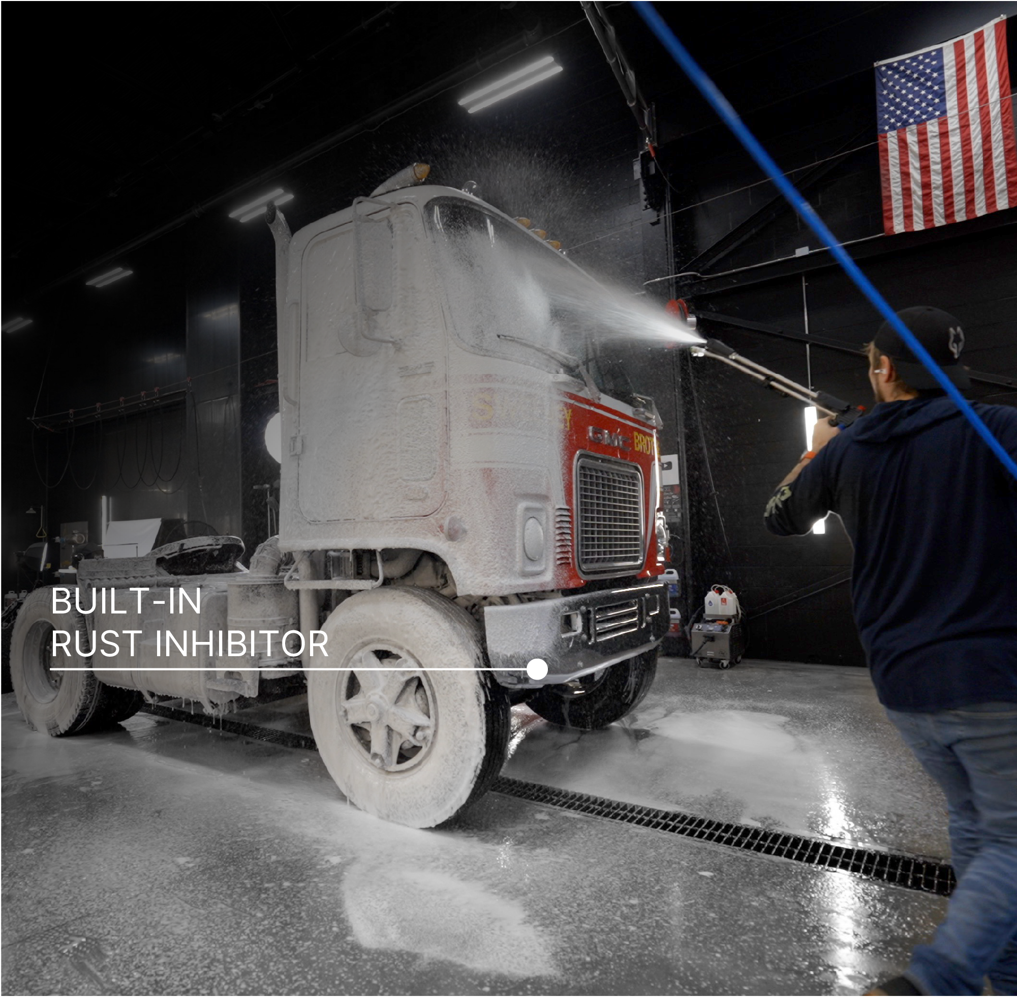 A semi-truck cab is being washed with a high-pressure water spray inside a wash bay. The truck is covered in white foam, and a person on the right is spraying it. There is text overlaid on the left side of the screen reading 'BUILT-IN RUST INHIBITOR.'