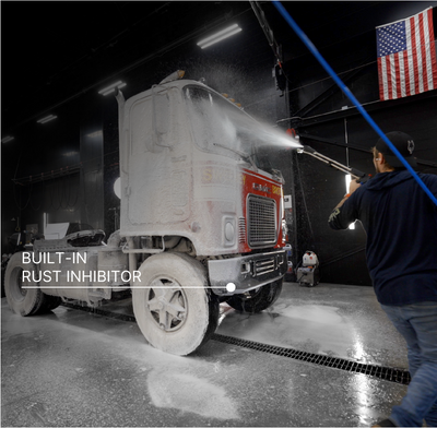 A semi-truck cab is being washed with a high-pressure water spray inside a wash bay. The truck is covered in white foam, and a person on the right is spraying it. There is text overlaid on the left side of the screen reading 'BUILT-IN RUST INHIBITOR.'
