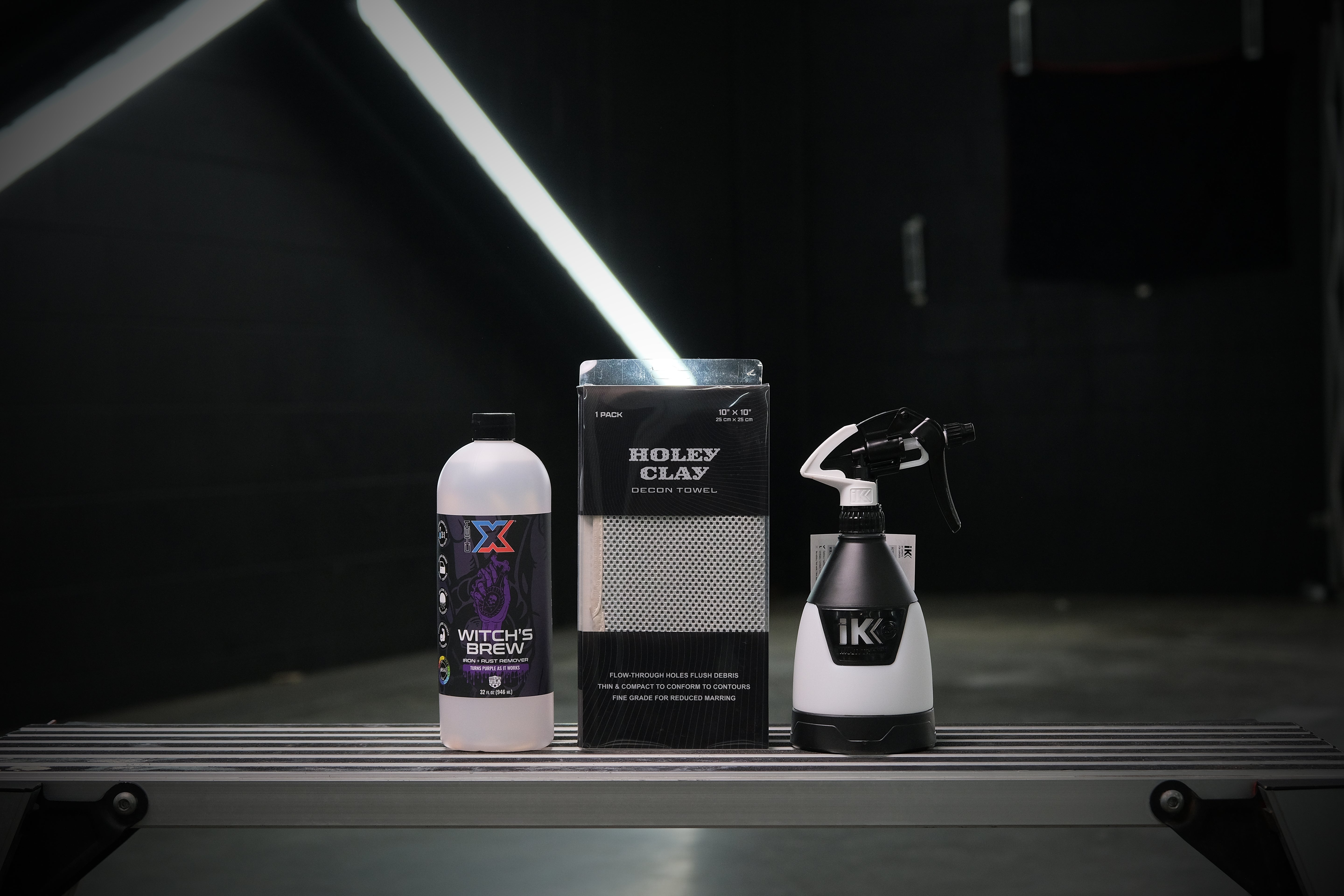 A grey towel with holes in a clear box called 'Holey Clay Decon Towel' next to a 32 oz bottle of clear iron and rust remover called 'Witch's Brew,' and a black and white trigger sprayer with the letters 'iK' on the front lined up on a workbench in a dark wash bay setting with fluorescent lighting in the background.