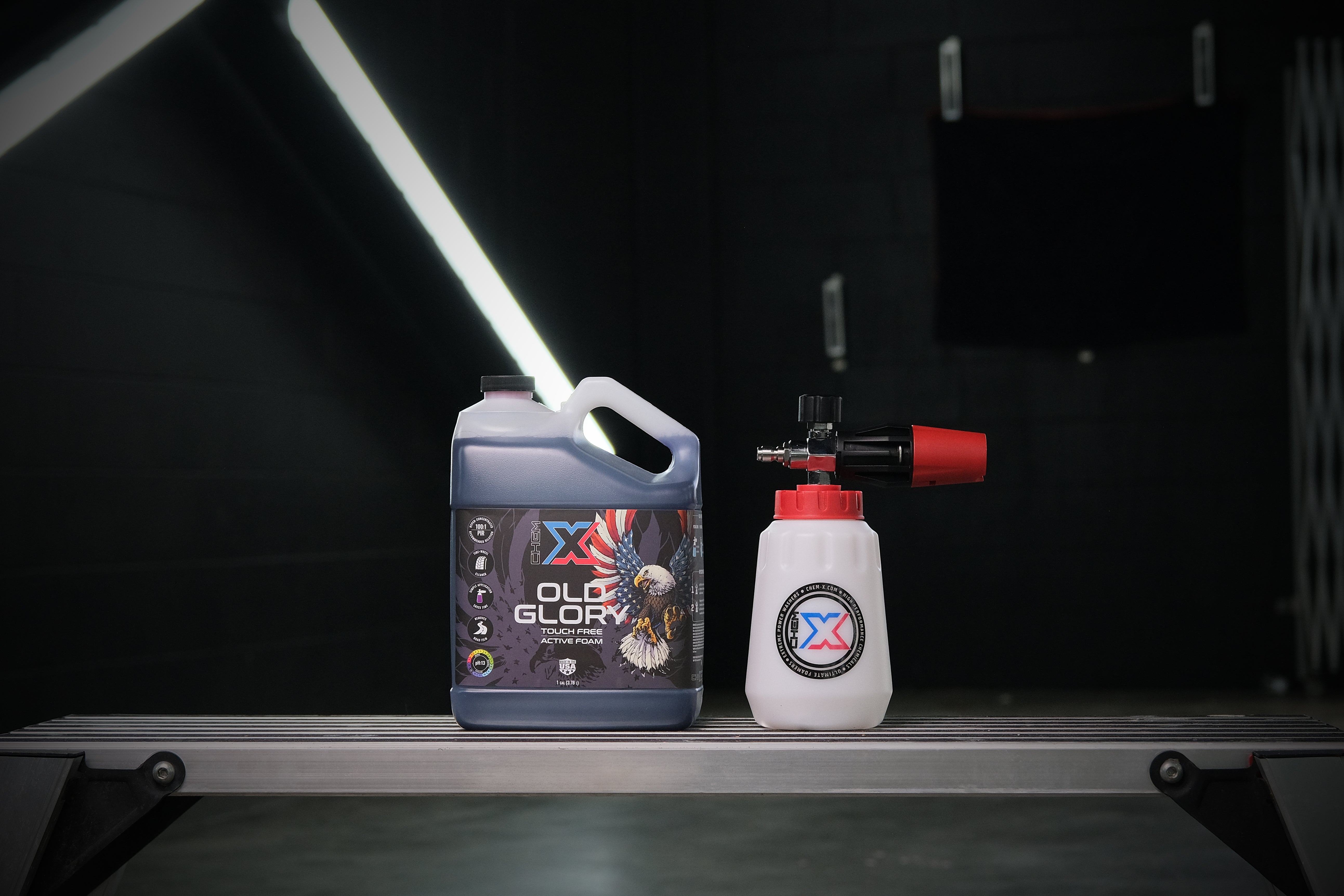 One gallon bottle of black touch free active foam vehicle wash called 'Old Glory' next to a foam cannon with a red top on a workbench in a wash bay setting.