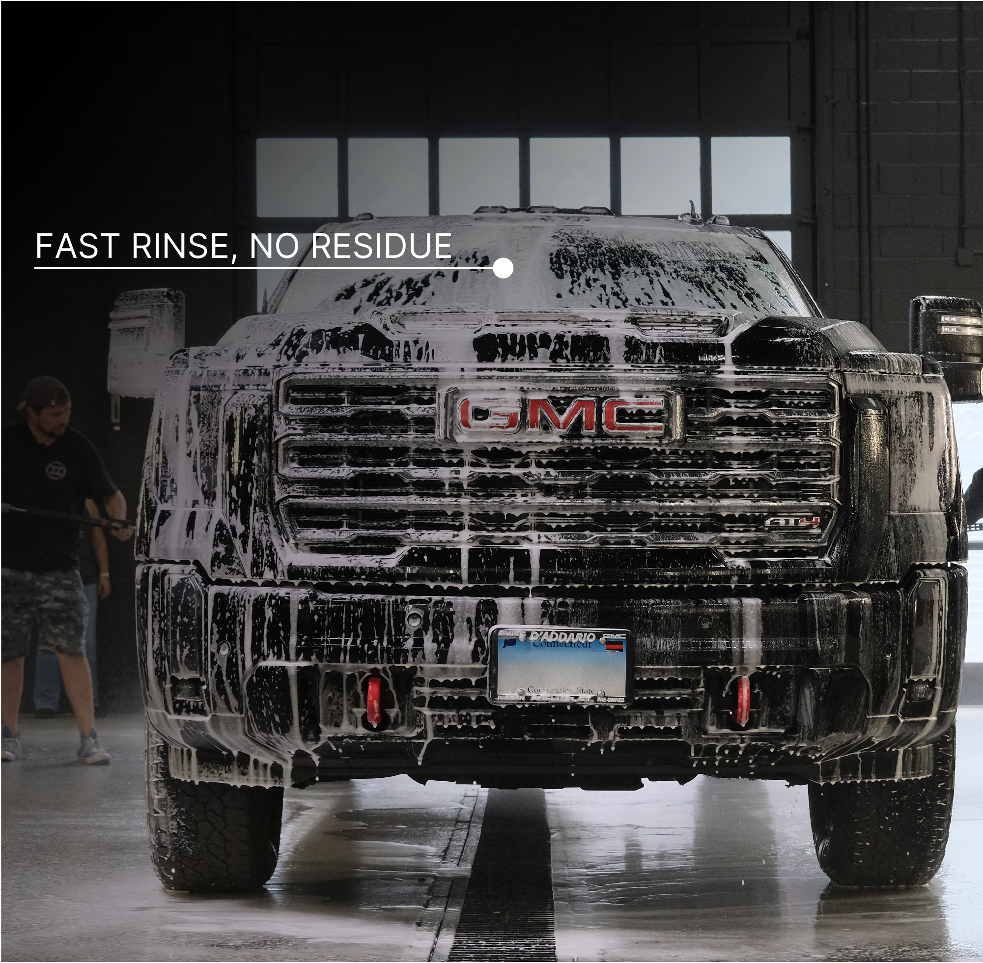 A black GMC pickup truck covered in white soap foam, photographed from the front in a wash bay. White text to the left reads 'FAST RINSE, NO RESIDUE.'