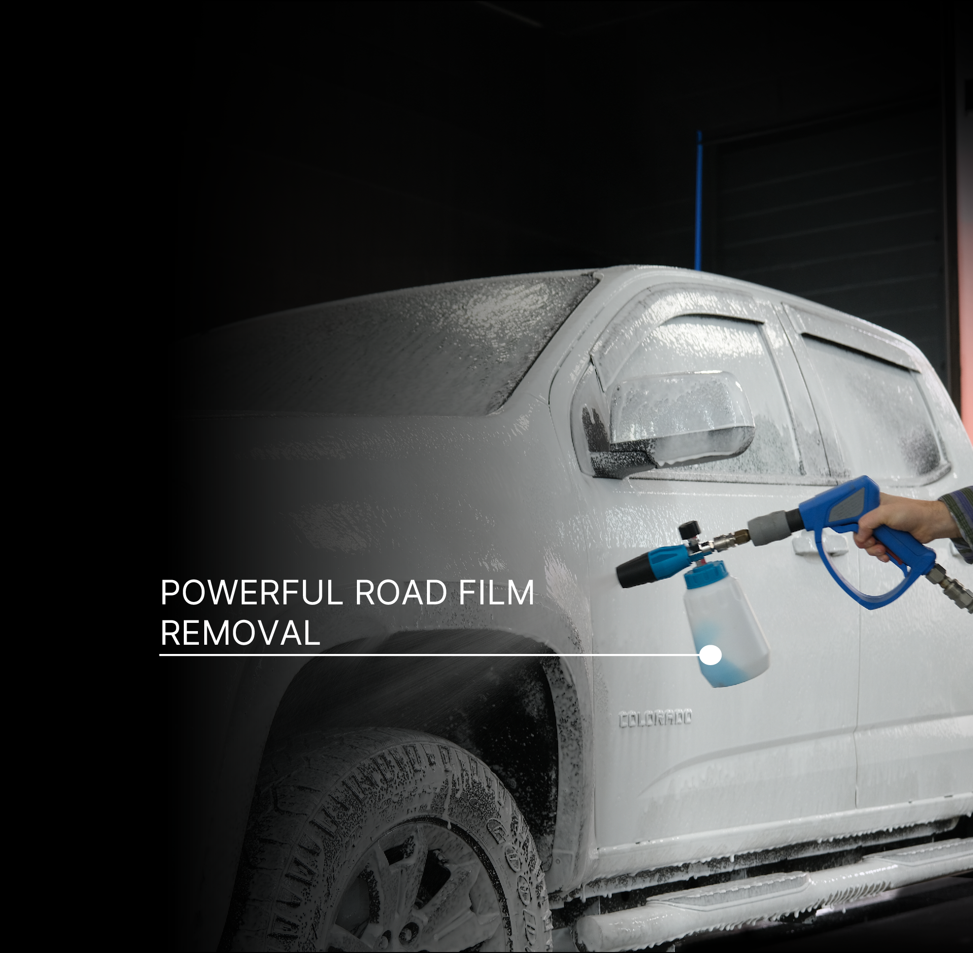 A hand holds a blue and black foam cannon sprayer, applying foam to a white pickup truck covered in road film, with the text 'POWERFUL ROAD FILM REMOVAL' on a dark background.