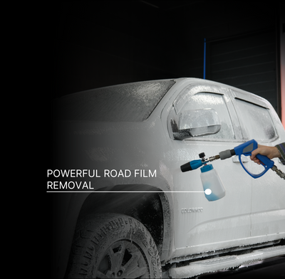 A hand holds a blue and black foam cannon sprayer, applying foam to a white pickup truck covered in road film, with the text 'POWERFUL ROAD FILM REMOVAL' on a dark background.
