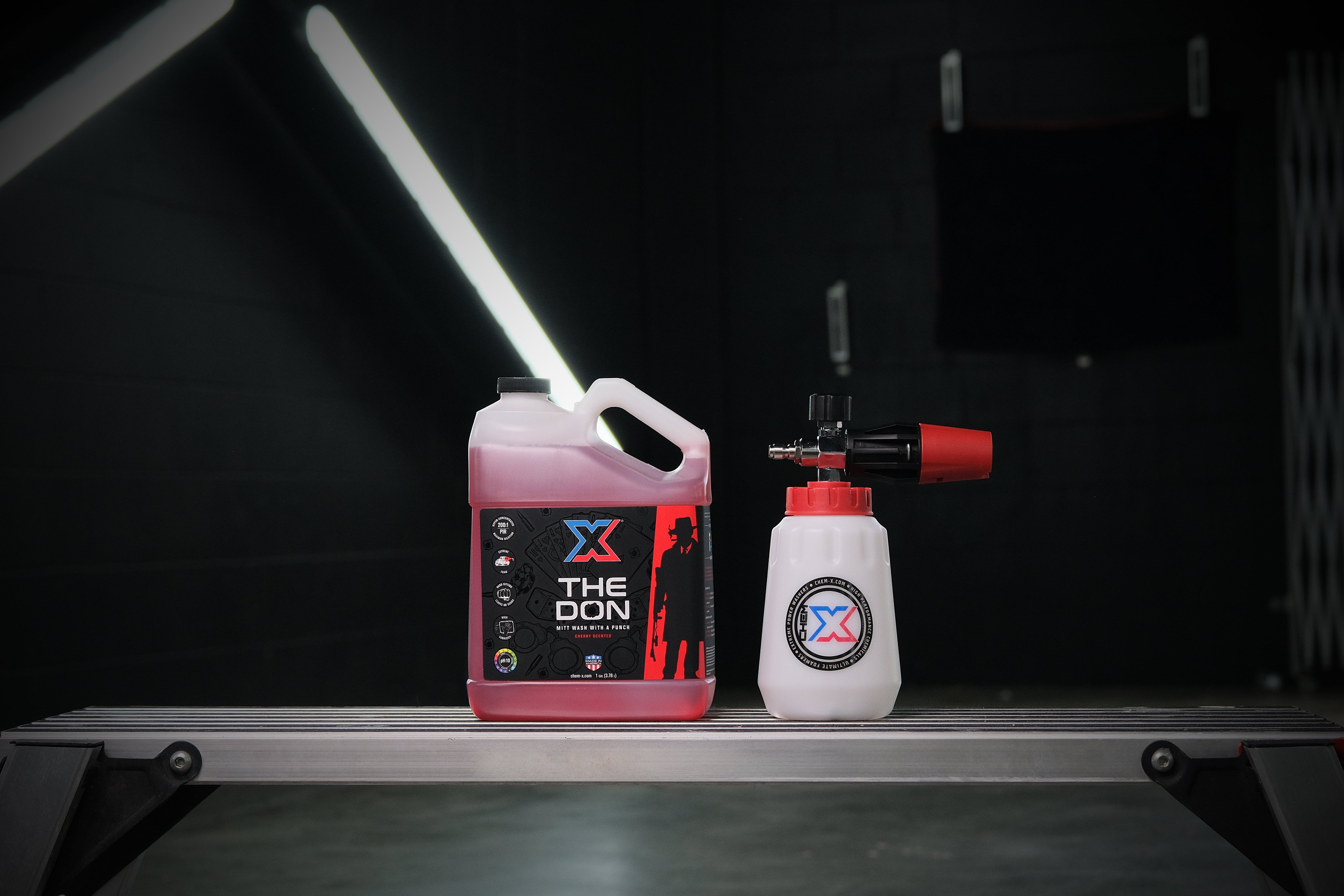 One gallon bottle of red mitt wash called 'The Don' next to a foam cannon with a red top sitting on a workbench in a dark wash bay setting with fluorescent lights in the back.